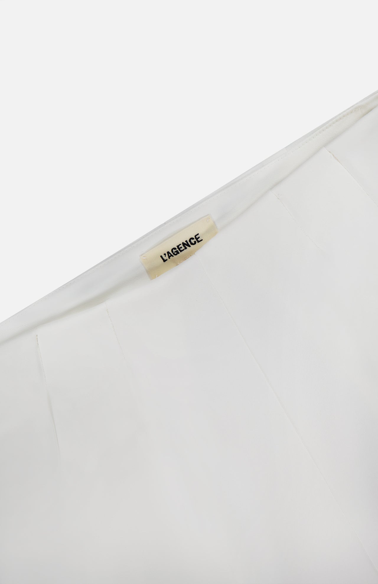 Close-up of a white garment with a beige L’AGENCE label sewn into the collar. Style it with the Novalynn Cropped Pleated Pant by L'AGENCE for a chic look. The background is plain white.