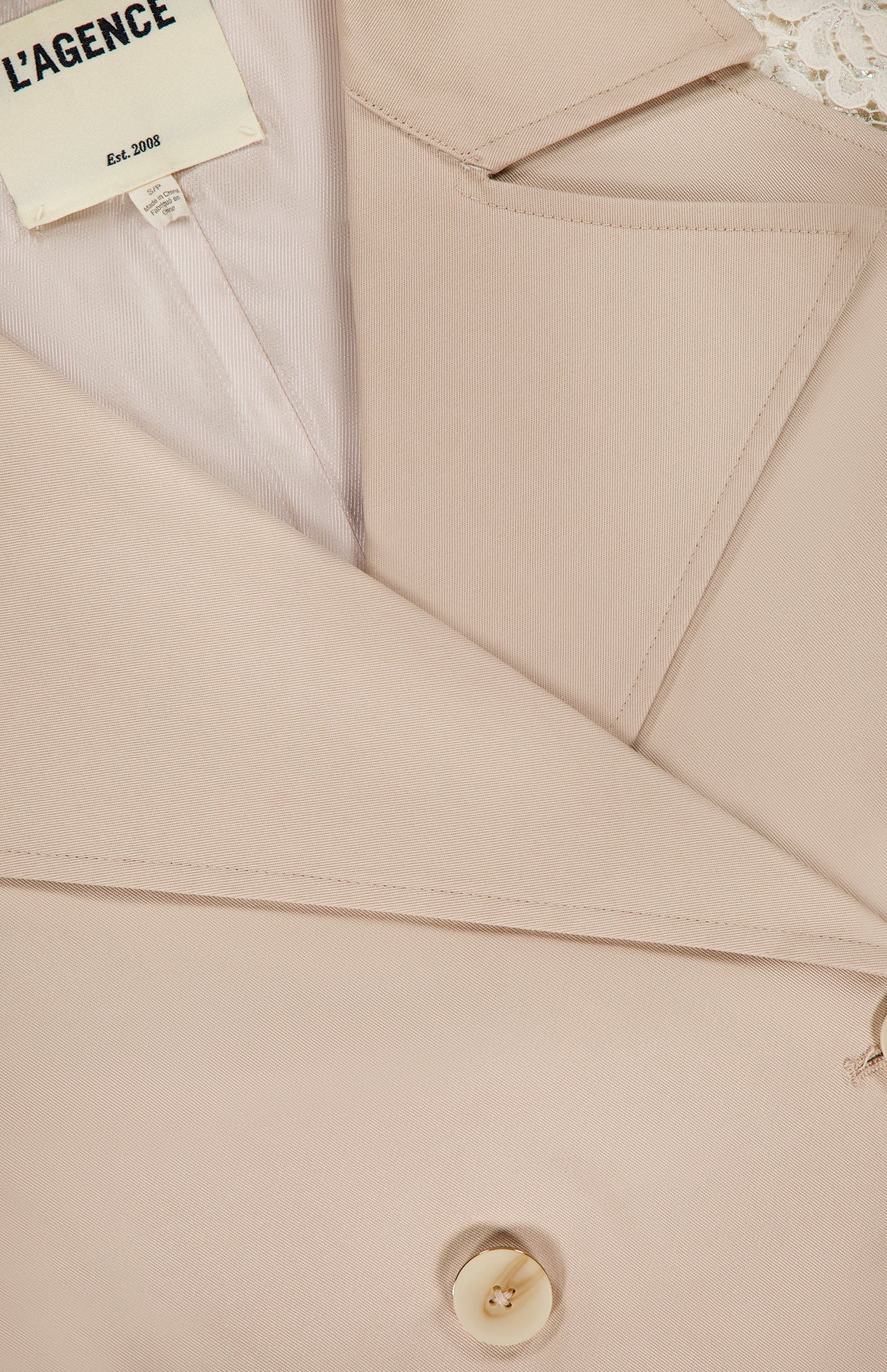 Close-up of the Novelle Lace Sleeve Trench Coat by L'AGENCE, featuring beige fabric, button, lapel, pocket, and a visible "L'AGENCE Est. 2008" label. White floral lace sleeve detail peeks from the top right corner.