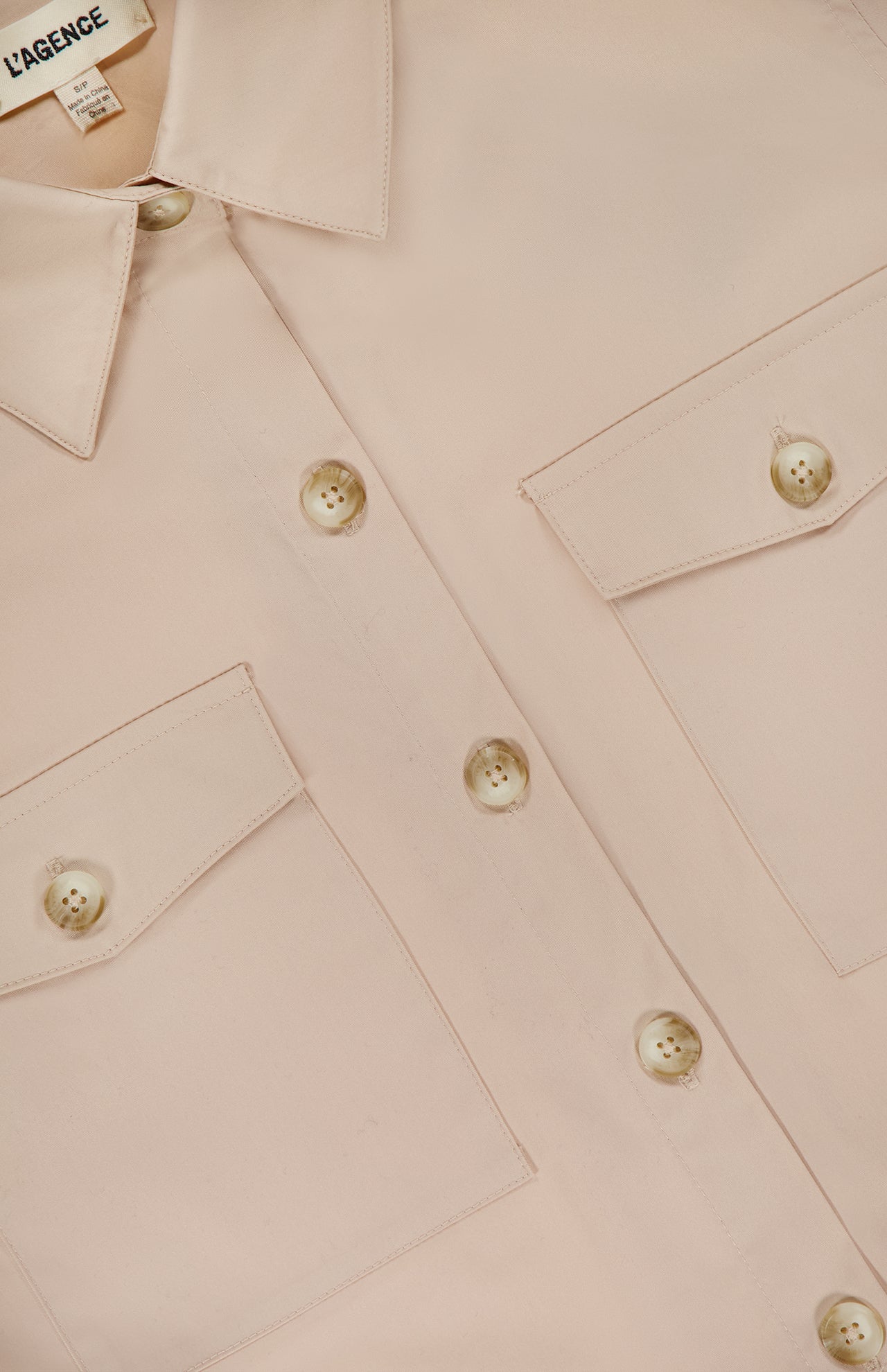 Close-up of the Pearson Military Pocket Blouse by L'AGENCE in light pink poplin, featuring two front chest pockets and beige buttons. The partially visible L'AGENCE label adds a chic touch to this versatile button-down top.