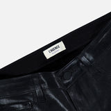 Close-up of the waistband and upper portion of the Petite Kendra High Rise Crop Flare in Noir by L’AGENCE, featuring shiny black coated denim with belt loops, pockets, a button closure, and a white L’AGENCE label. The background is plain and light-colored.