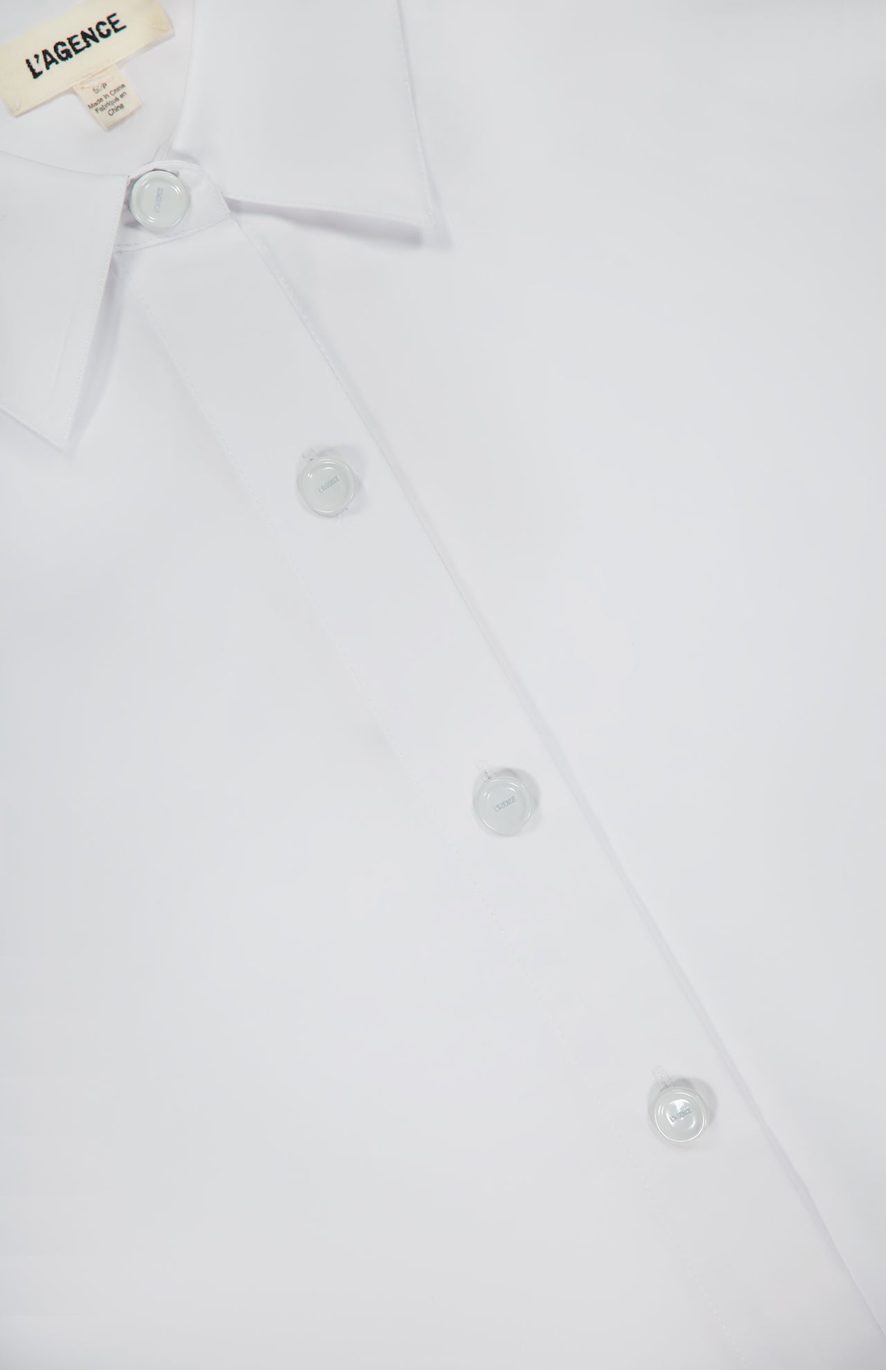 Close-up of the Rylie Relaxed Dress Shirt by L’AGENCE, featuring white organic cotton, a pointed collar, four visible buttons, and an inner collar label that reads "L'AGENCE" in black.