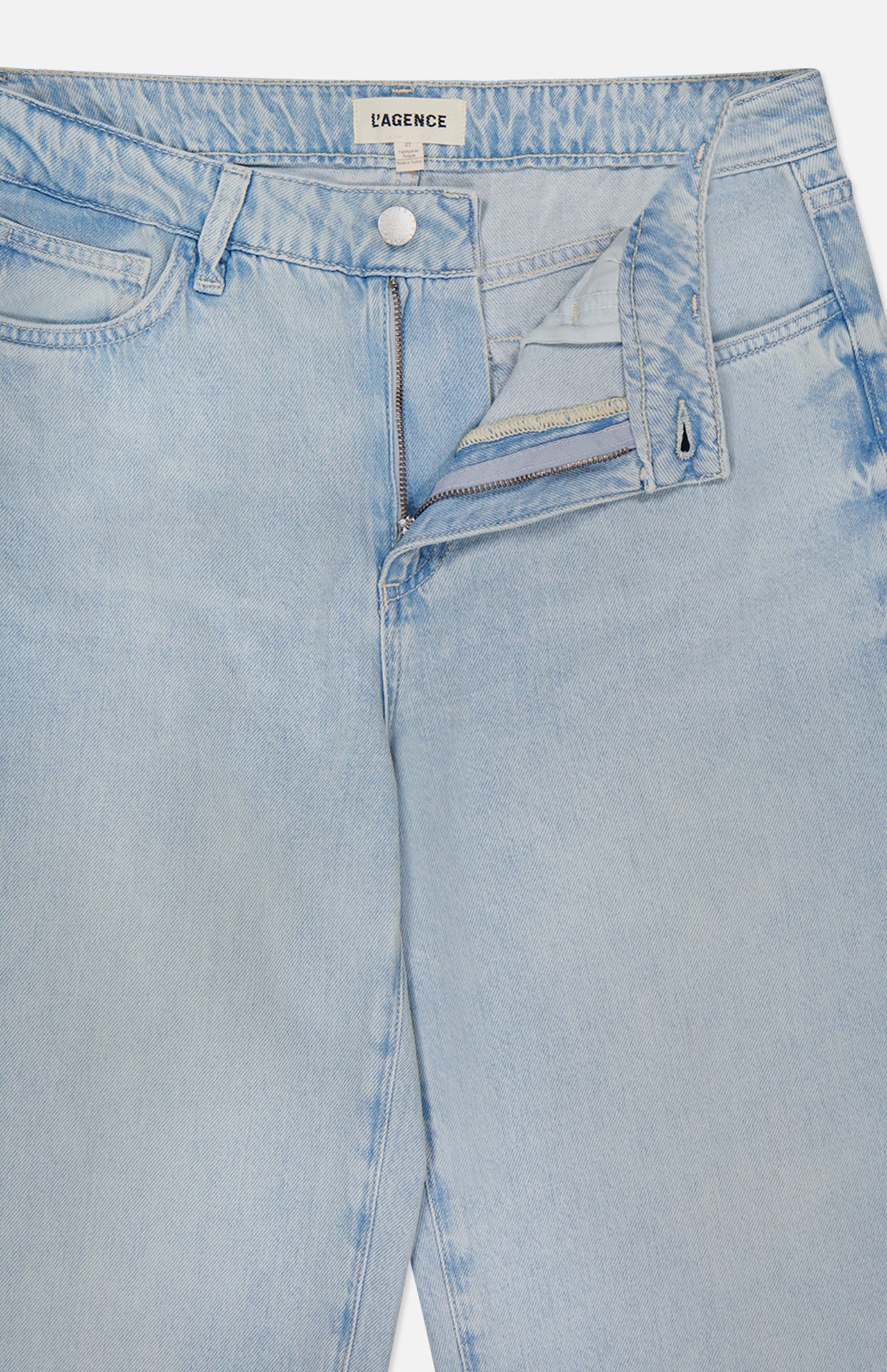 The Scottie High Rise Wide Leg Jean in Lago by L'AGENCE features light blue denim with a partially open zipper and button fly, shown lying flat on a white surface. The waistband label displays the L'AGENCE brand name.