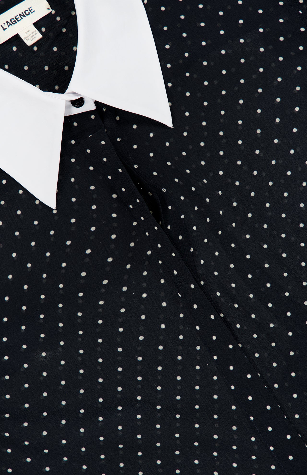 Close-up of the Sona Poplin Collar Blouse by L’AGENCE, featuring a black base adorned with small white polka dots and finished with a crisp white collar for an effortlessly chic look. The partially visible label at the neckline adds a subtle touch to this office-appropriate top.