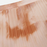 Close-up of the Torin Pleated Maxi Skirt by L'AGENCE in light peach, showcasing subtle brown and pink frappe tie-dye. The soft fabric features evenly spaced vertical pleats and a smooth, slightly glossy finish.