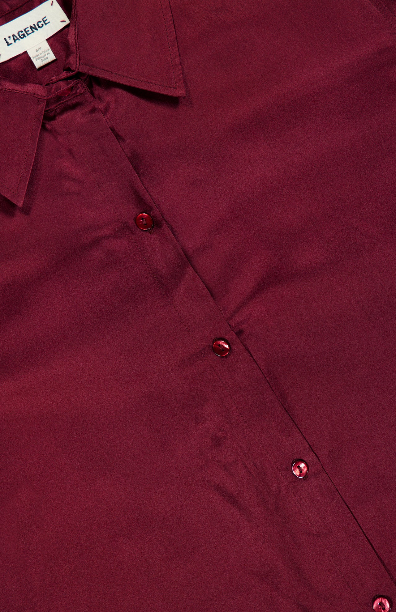 Close-up of a shiny merlot red Tyler Long Sleeve Silk Blouse, featuring a pointed collar and a visible L’AGENCE label in black text on a white background. This elegant blouse by L’AGENCE is crafted from luxurious silk, showcases long sleeves, a classic button-down design, and an unmistakable sheen.