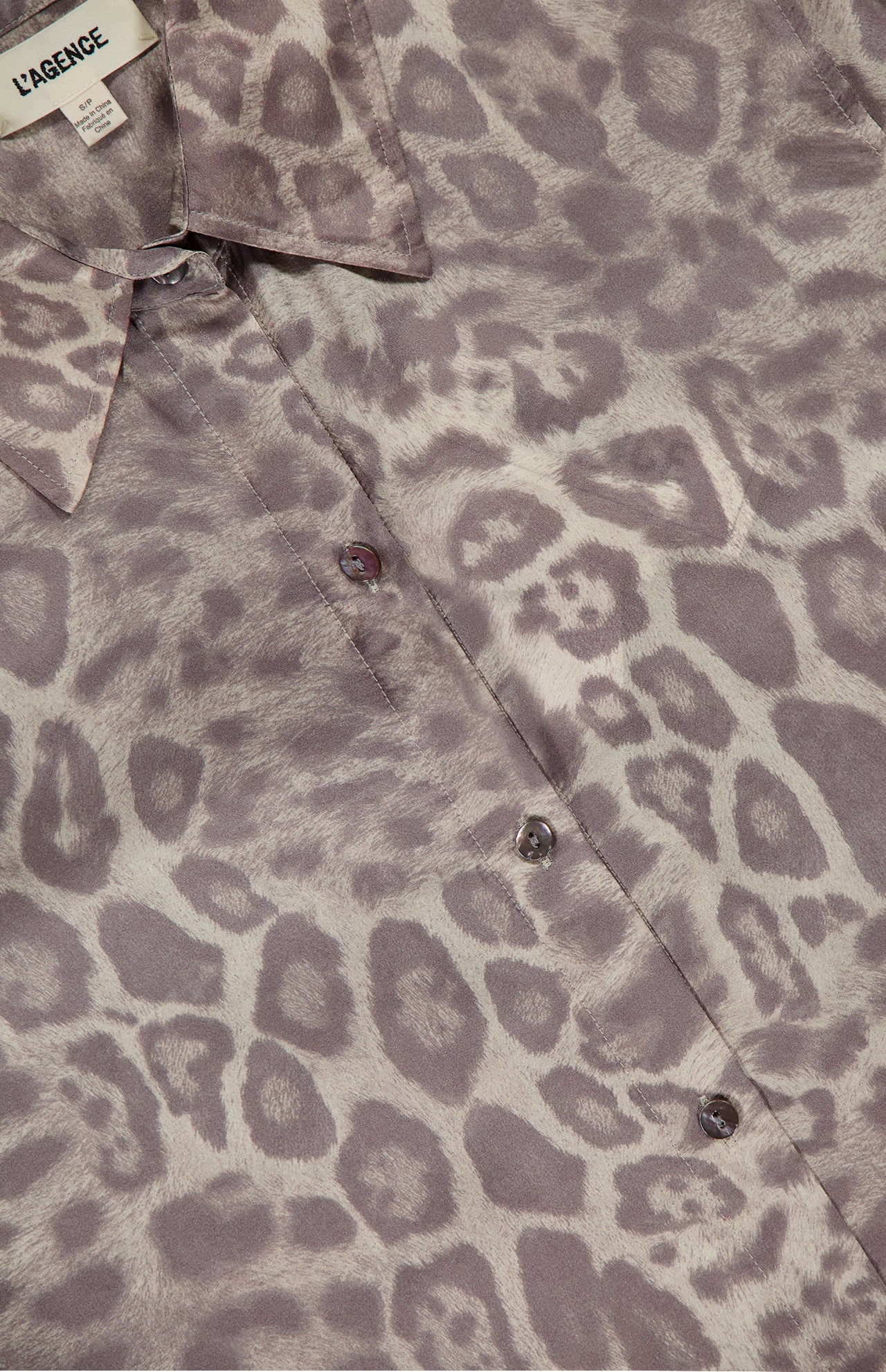 Close-up of the L'AGENCE Tyler Long Sleeve Silk Leopard Print Blouse, showcasing its pale purple and grey leopard pattern, button-down front, collar, and signature label at the neckline.