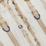 Close-up of the L’AGENCE Tyler Long Sleeve Belt Print Blouse, a white silk button-down top featuring an elegant pattern of beige belts, blue rings, and gold buckles. The brand’s label is visible on the collar of this sophisticated women’s blouse.