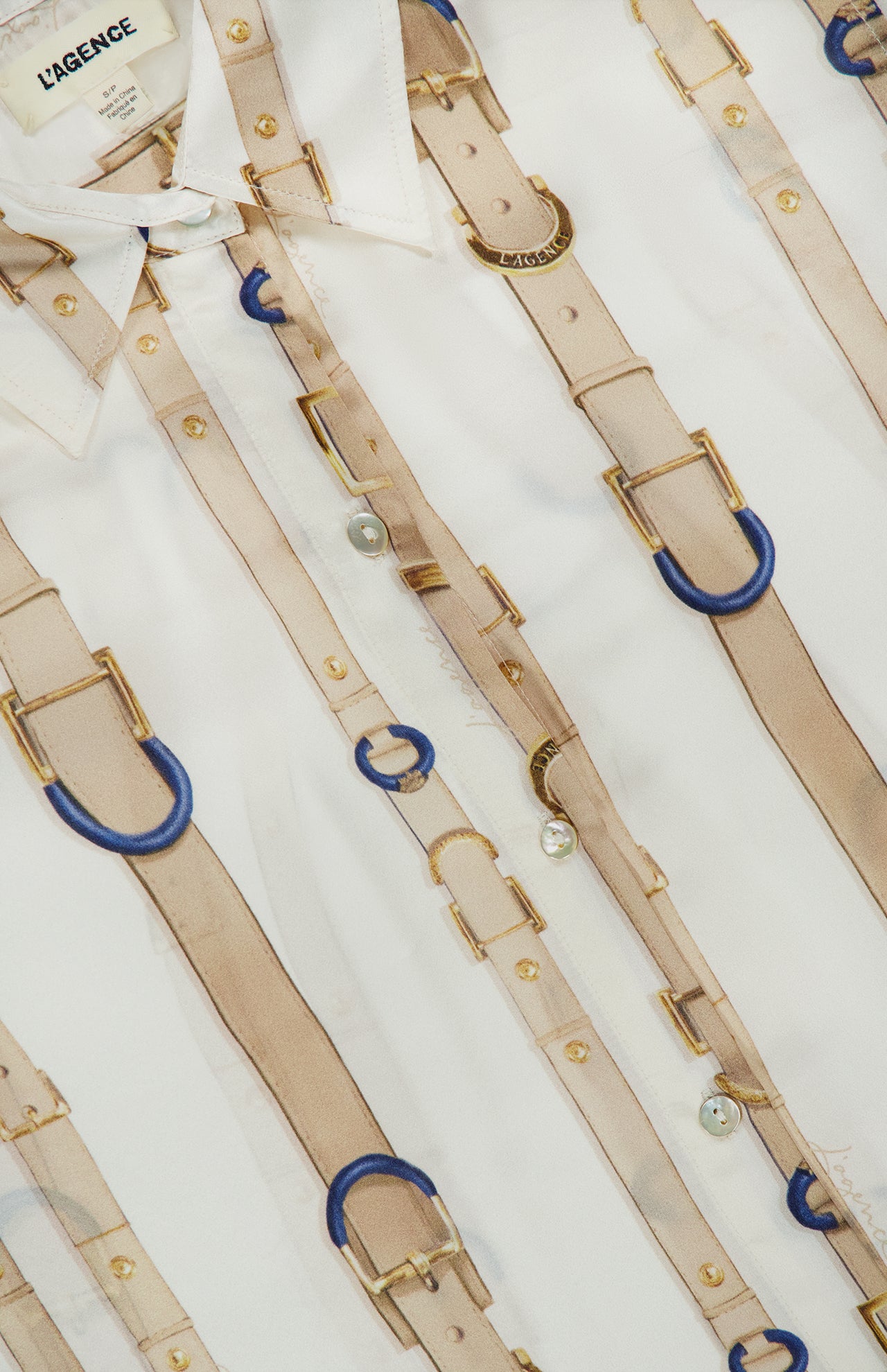 Close-up of the L’AGENCE Tyler Long Sleeve Belt Print Blouse, a white silk button-down top featuring an elegant pattern of beige belts, blue rings, and gold buckles. The brand’s label is visible on the collar of this sophisticated women’s blouse.