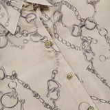 Close-up of the L'AGENCE Tyler Long Sleeve Chain Print Blouse in beige silk, featuring a tailored fit, collar, horsebit chain and jewelry print, pearly buttons, subtle sheen, and visible L'AGENCE tag on the shirt.