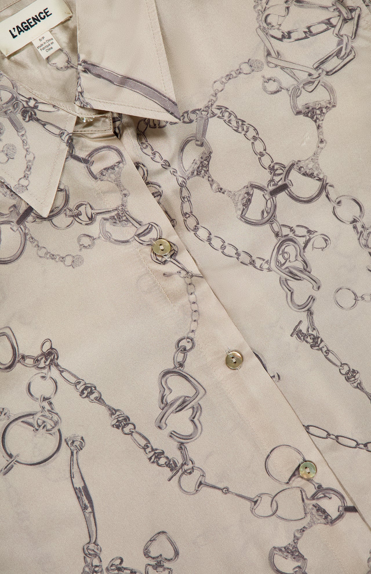 Close-up of the L'AGENCE Tyler Long Sleeve Chain Print Blouse in beige silk, featuring a tailored fit, collar, horsebit chain and jewelry print, pearly buttons, subtle sheen, and visible L'AGENCE tag on the shirt.