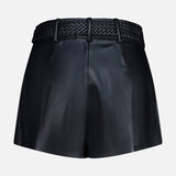 The Virelle Braided Waistband Short by L’AGENCE features black vegan leather with a high waist, front pleats, side pockets, and a braided waistband threaded through belt loops, displayed against a plain white background.