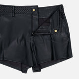The Virelle Braided Waistband Short by L’AGENCE features black vegan leather, a braided waistband, belt loops, a gold button, and a zipper fly. Styled partially open against a plain light background, these shorts exude a chic, western-inspired vibe.