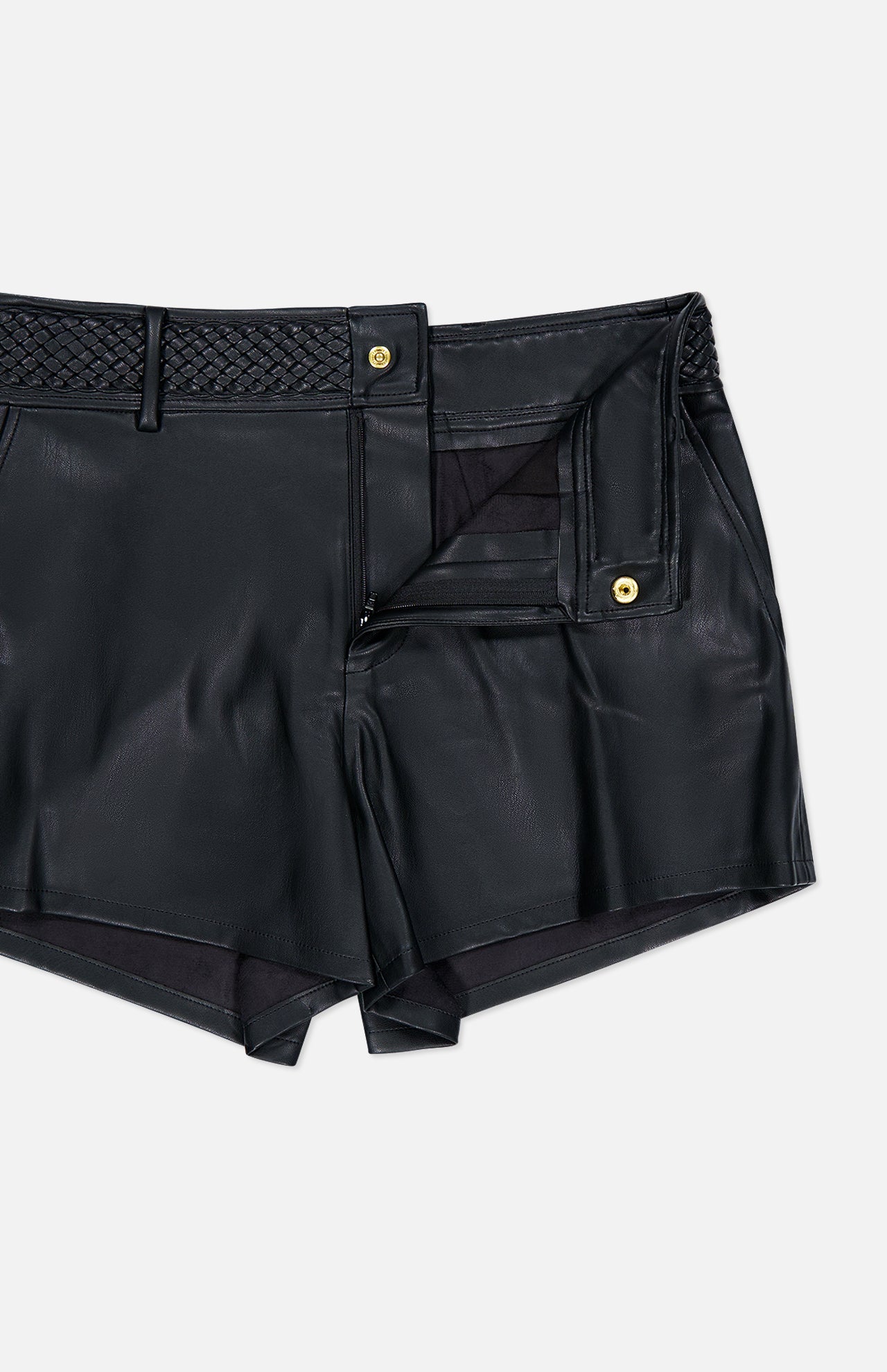 The Virelle Braided Waistband Short by L’AGENCE features black vegan leather, a braided waistband, belt loops, a gold button, and a zipper fly. Styled partially open against a plain light background, these shorts exude a chic, western-inspired vibe.