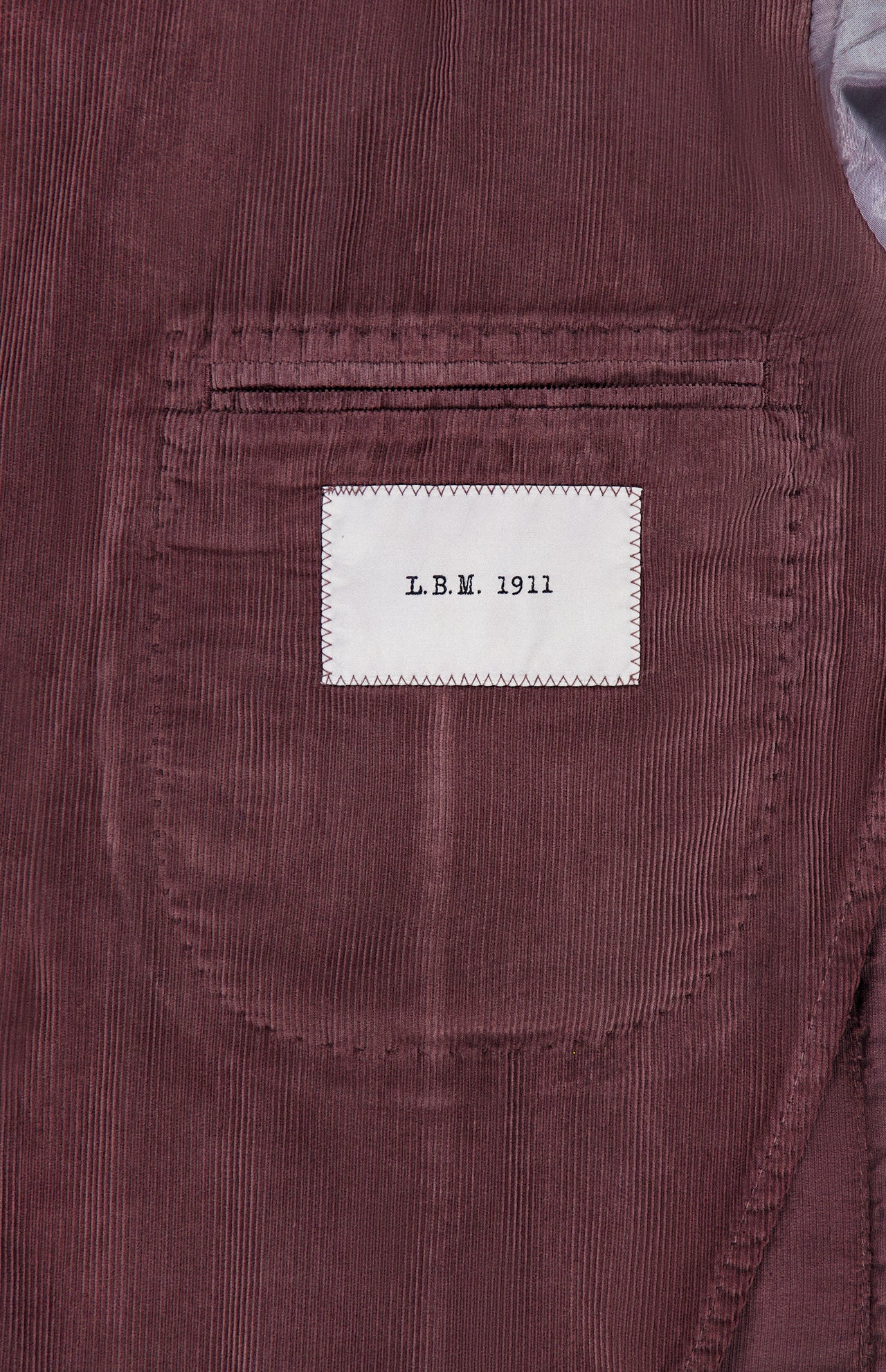 A close-up of the pocket on the Corduroy Single Breasted Sportcoat by L.B.M. showcases its rich maroon corduroy texture and a distinctive white label featuring “L.B.M. 1911” in black lettering, emphasizing the brand’s refined craftsmanship in this classic men’s jacket.