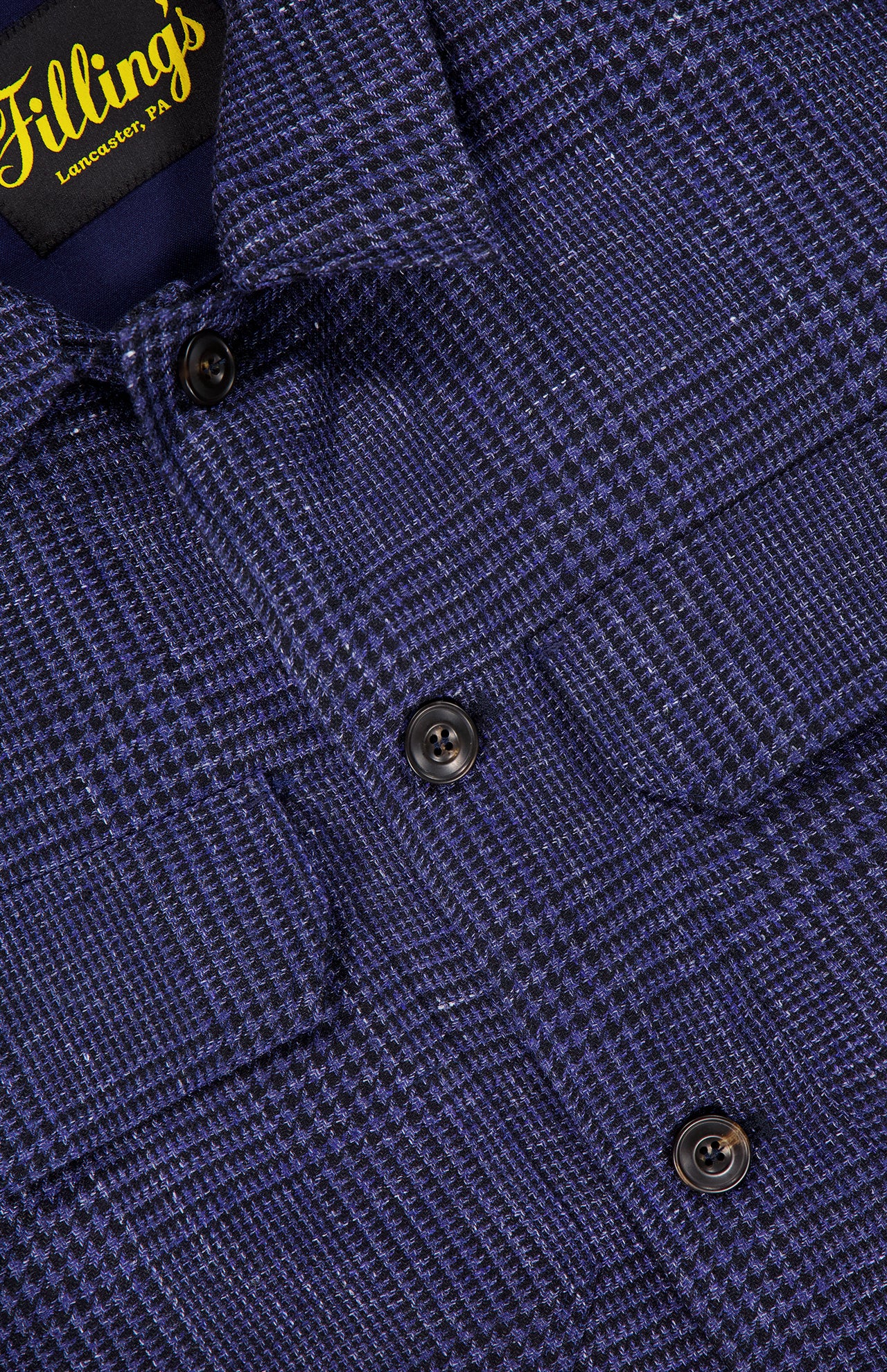 Close-up of the Glen Plaid Overshirt by L.B.M., showcasing a navy plaid pattern with black buttons and two chest pockets. The soft texture suggests the premium quality of this men's button-front shirt. The Yellow Fillings Lancaster, PA label is visible on the inside collar.