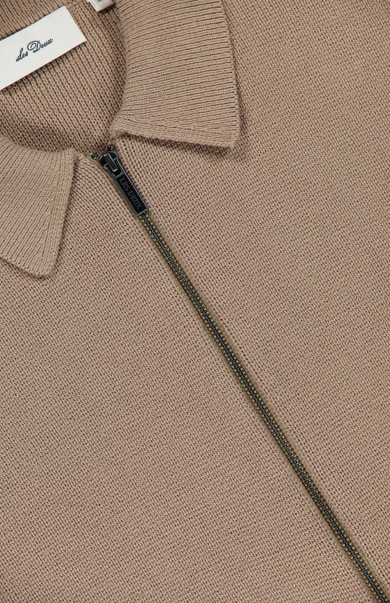 Close-up of the beige Fischer Cotton Milano Zip Overshirt by LES DEUX, featuring a structured knit, collar, and partially unzipped dark metal zipper. A white brand tag is visible inside the collar, showcasing this versatile piece.
