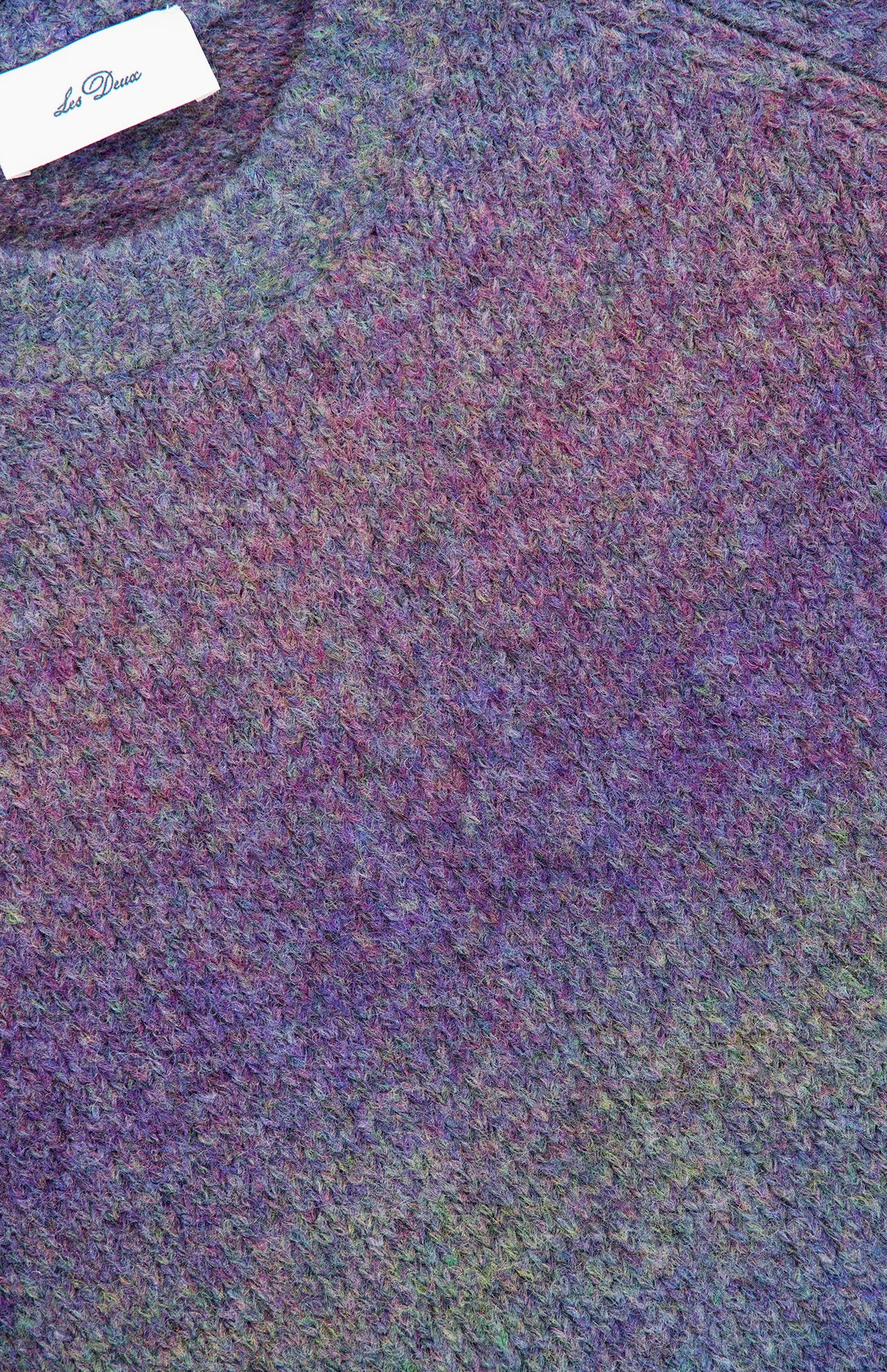 Close-up of the LES DEUX Gradient Roundneck Knit Sweater, showcasing its textured knit and gradient-dyed shades of spiced plum, green, and blue. A white clothing tag with text is visible near the collar.