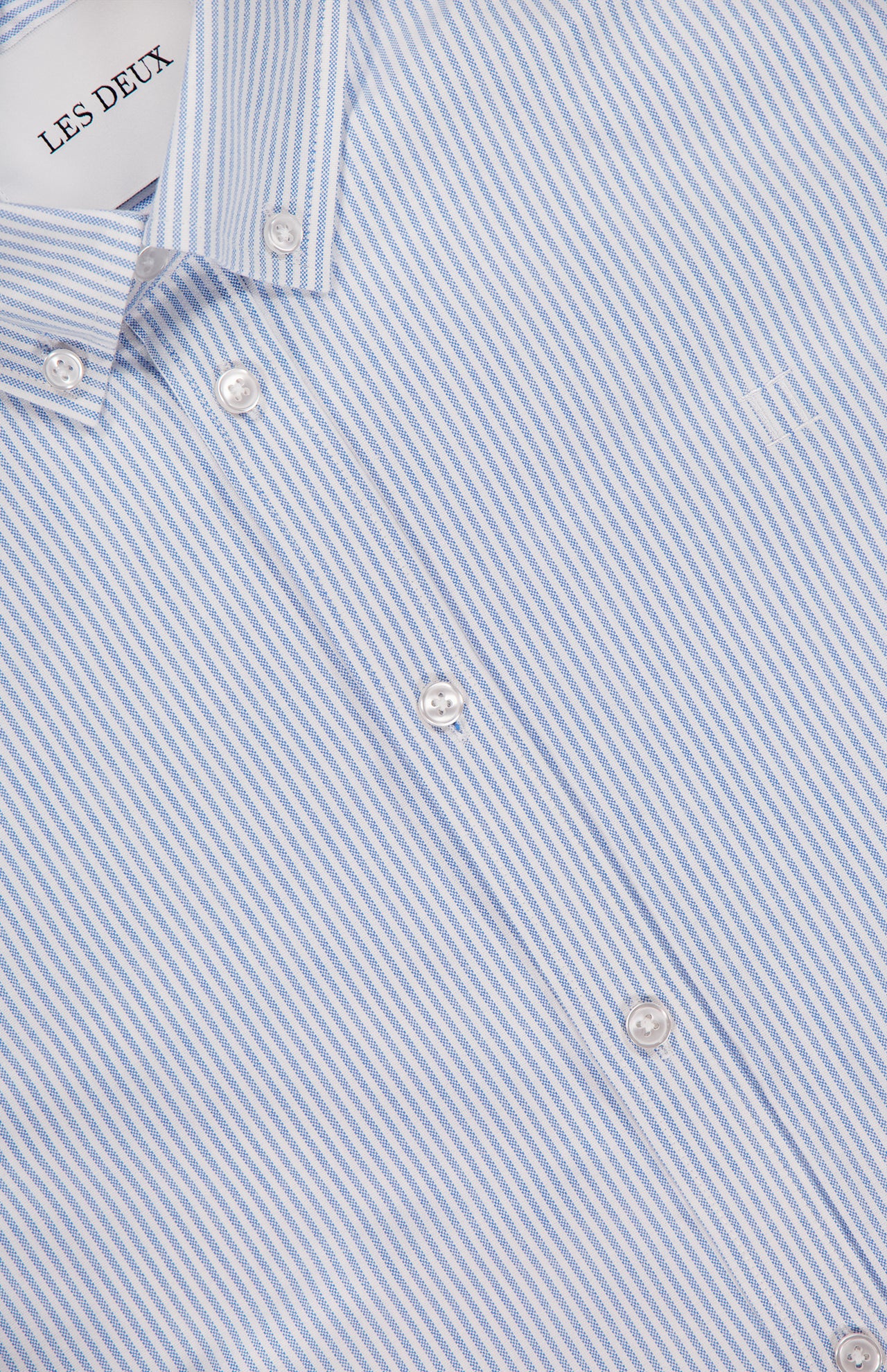 A close-up of the LES DEUX Konrad Striped Oxford Shirt in light blue and white shows white buttons, a small embroidered logo on the chest, and classic stripes.