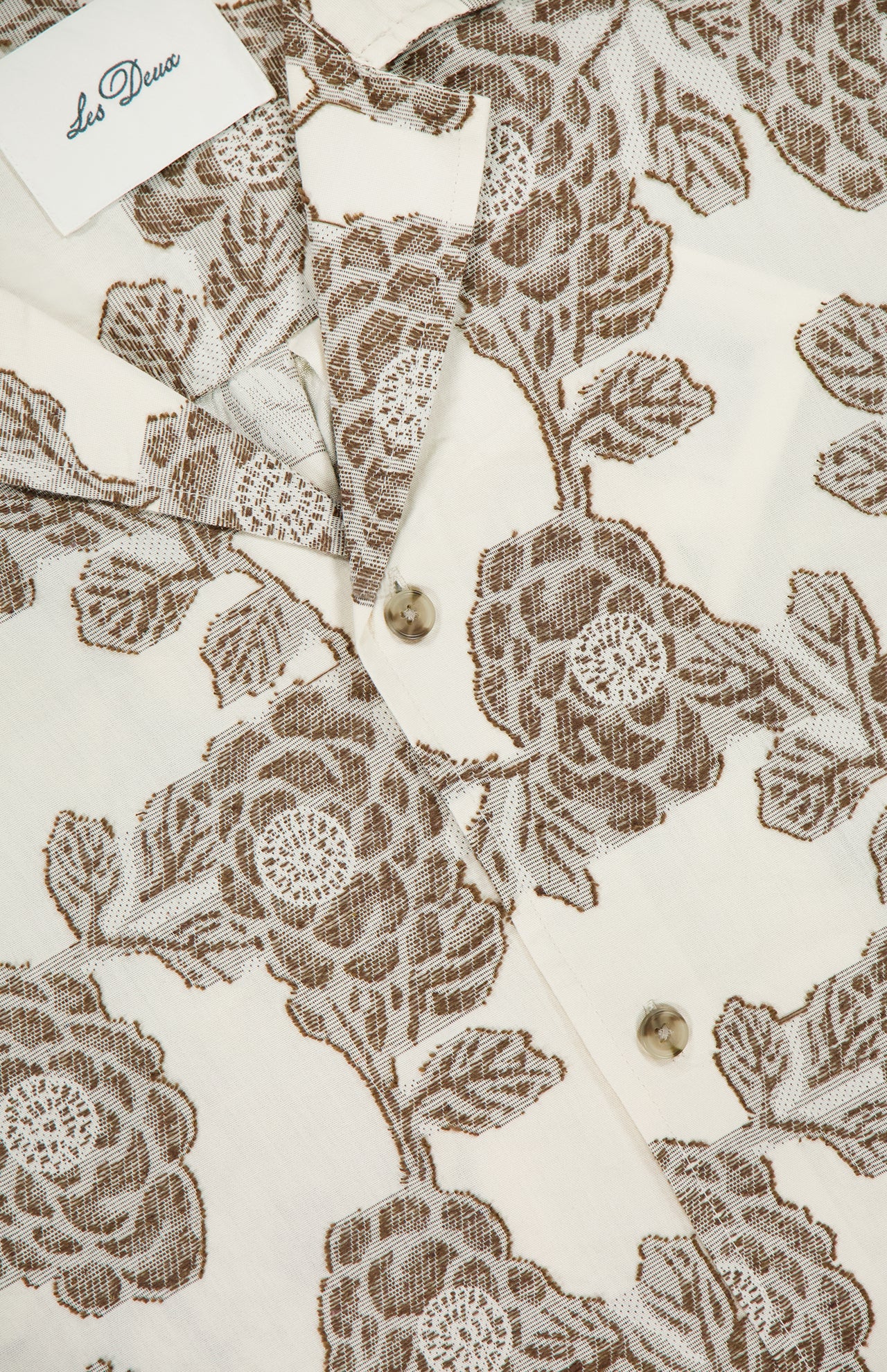 A close-up of the Lesley Flower Jacquard Short Sleeve Shirt by LES DEUX shows its relaxed fit, cream floral jacquard fabric, beige buttons, and a white Les Deux label in the corner.