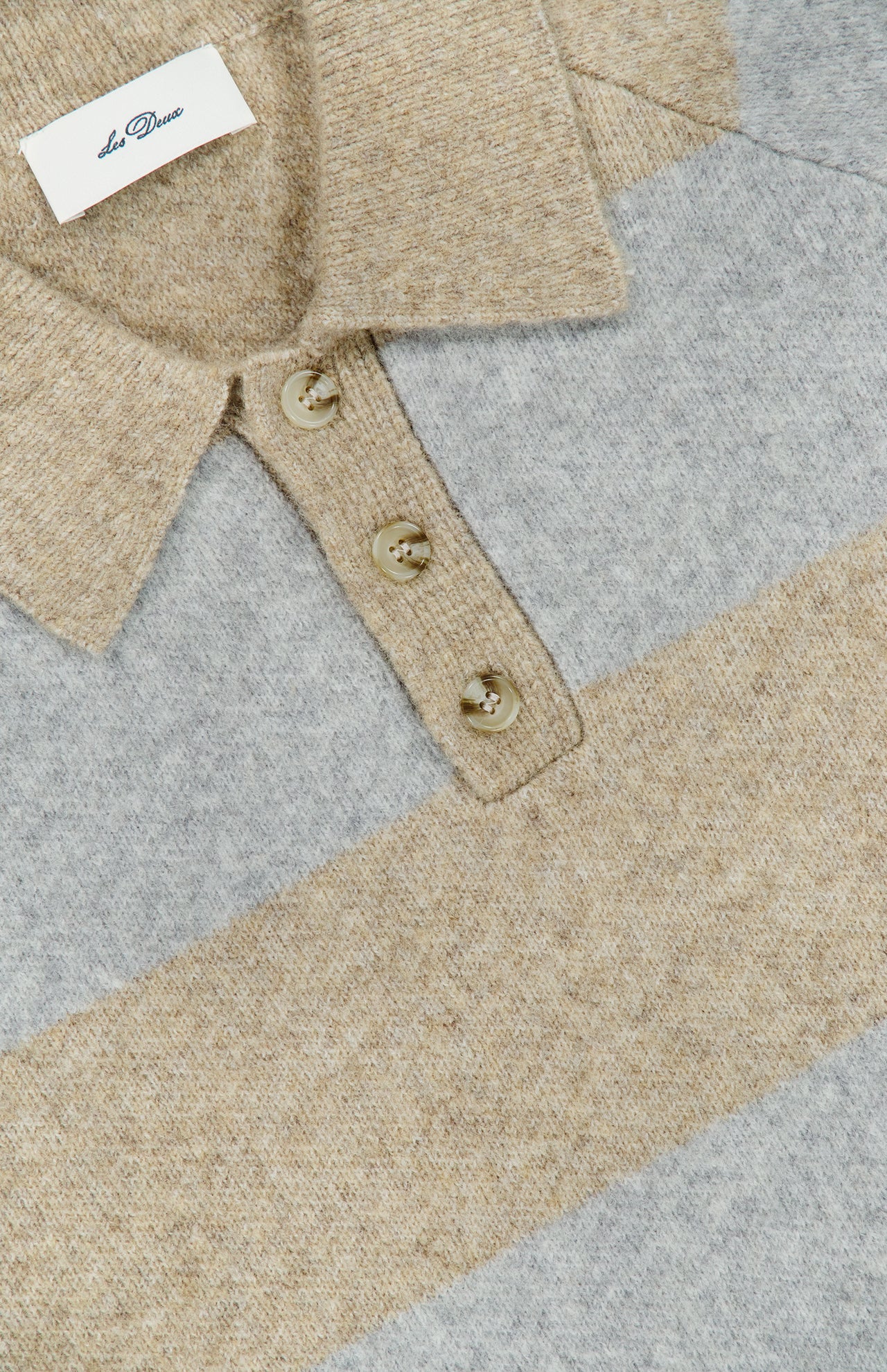 Close-up of the Striped Melange Knitted Polo Sweater by LES DEUX, featuring a collared neckline with three buttons and wide horizontal stripes in beige and gray melange. A white tag with text appears inside the collar, lending a classic, preppy finish to this stylish piece.