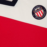 Close-up of the LES DEUX USA Football Jersey in ivory with a bold red diagonal stripe, navy collar, and a round Les Deux International Club patch with stars and stripes. A preppy urban clothing tag labeled LES DEUX is visible.
