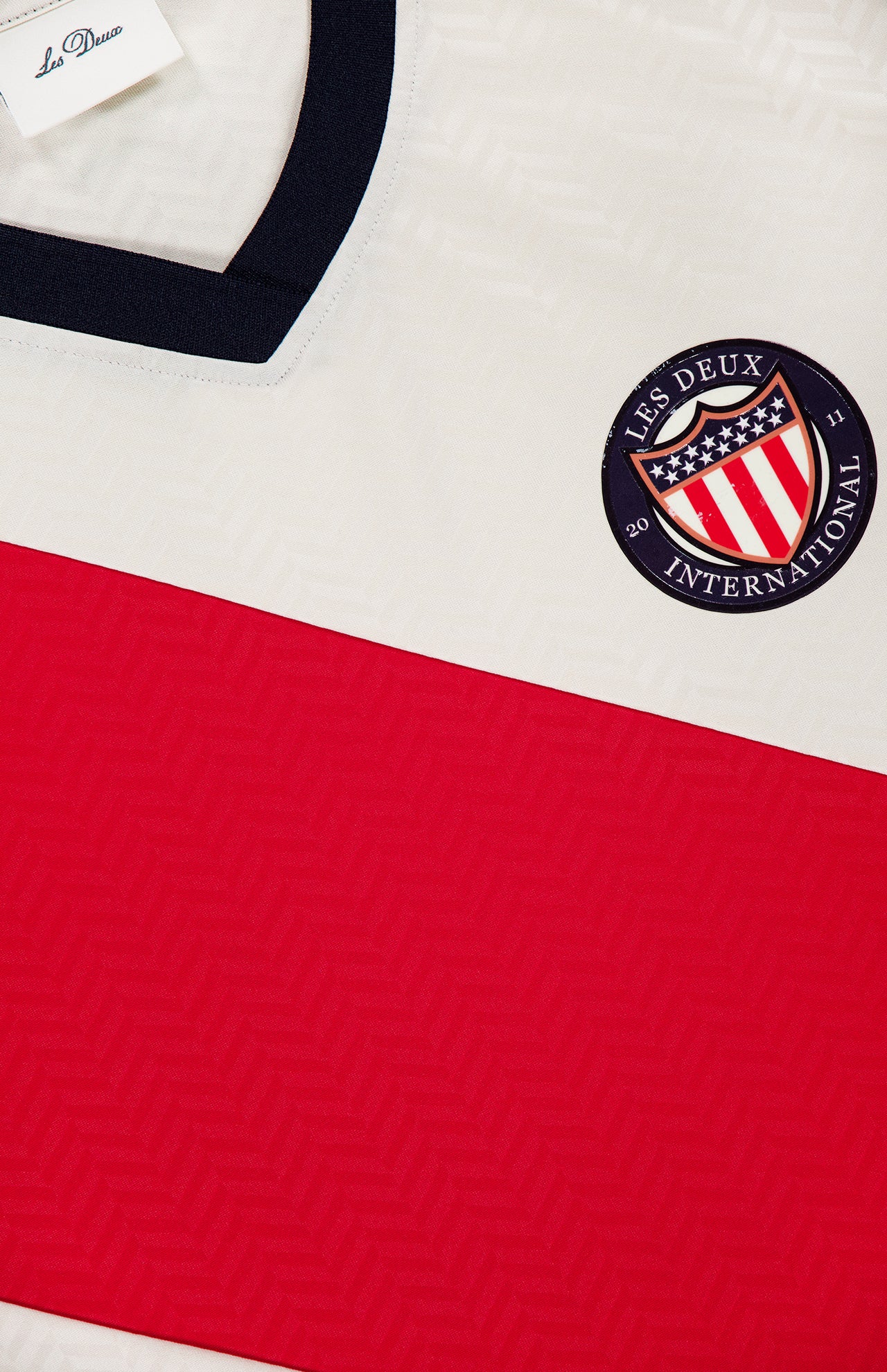 Close-up of the LES DEUX USA Football Jersey in ivory with a bold red diagonal stripe, navy collar, and a round Les Deux International Club patch with stars and stripes. A preppy urban clothing tag labeled LES DEUX is visible.