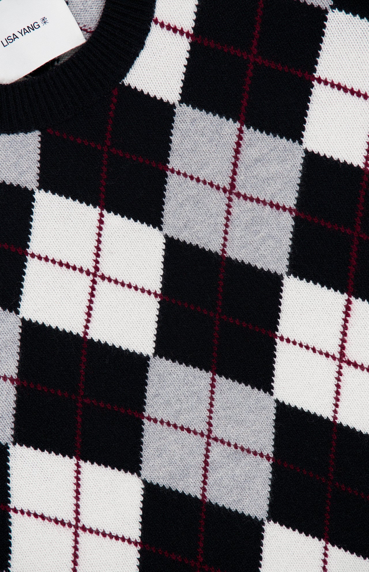 A close-up of the Antoni Cashmere Argyle Sweater by LISA YANG, showcasing its luxury black, white, and gray argyle pattern accented with thin red lines. Part of the garment’s label bearing the LISA YANG brand name is visible in the top left corner.
