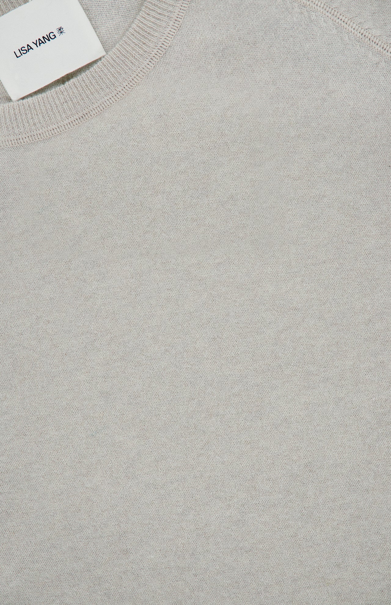 A close-up of the Cambrie Crewneck Sweater by LISA YANG showcases its light beige knit fabric, ribbed crew neckline, and a white inside-collar label featuring the LISA YANG brand in black text, all reflecting a Scandinavian aesthetic.