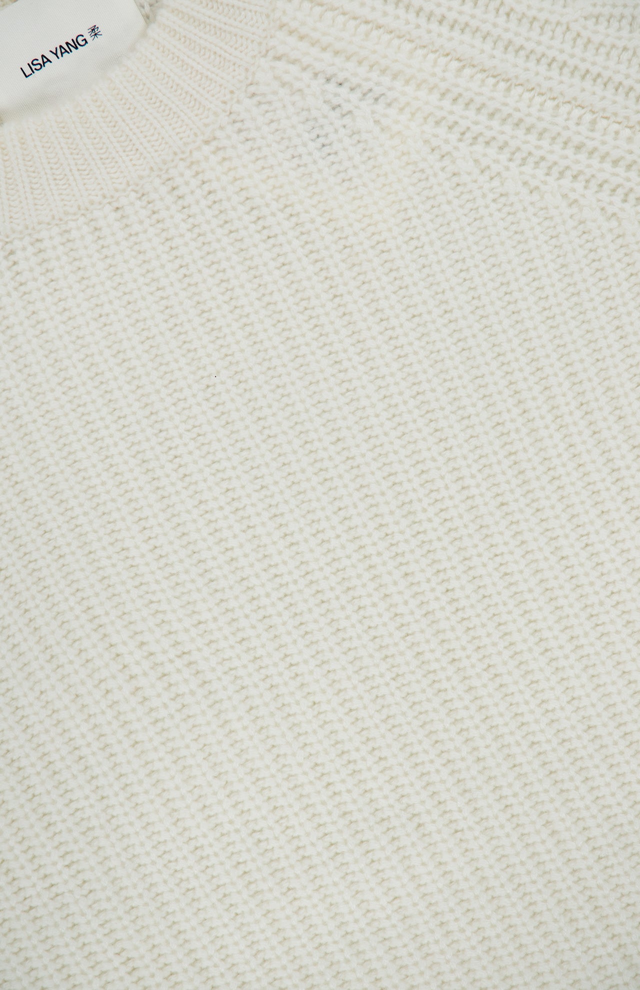 Close-up of the textured, off-white knit fabric of the Miki Cashmere Sweater. A small section of a label showing “LISA YANG,” the brand renowned for its pure cashmere pieces, appears at the top left corner.