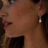 A woman with wavy brown hair and sun-kissed skin wears LOGAN HOLLOWELL’s Enigma Diamond Oracle Set Water Drop Earrings in 18k yellow gold, paired with a round gemstone necklace, gazing to the left.