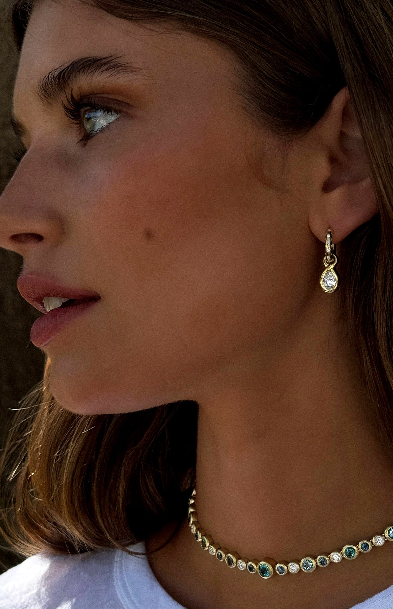 A woman with wavy brown hair and sun-kissed skin wears LOGAN HOLLOWELL’s Enigma Diamond Oracle Set Water Drop Earrings in 18k yellow gold, paired with a round gemstone necklace, gazing to the left.