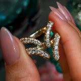 A hand with manicured nails holds three gold rings, including the stunning LOGAN HOLLOWELL French Pavé Diamond Tusk Ring in 14k yellow gold, encrusted with small diamonds, against a softly blurred, colorful background.
