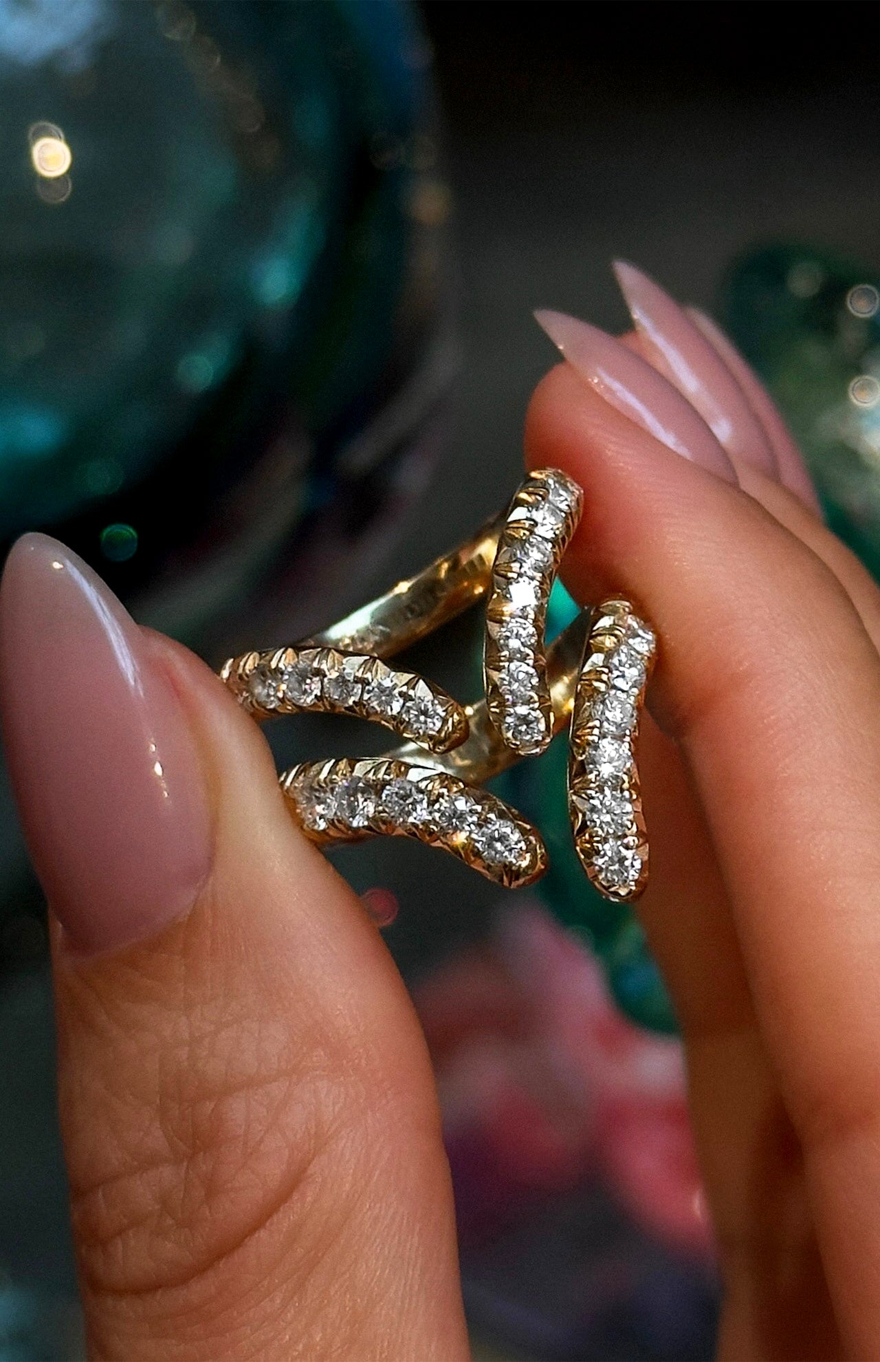 A hand with manicured nails holds three gold rings, including the stunning LOGAN HOLLOWELL French Pavé Diamond Tusk Ring in 14k yellow gold, encrusted with small diamonds, against a softly blurred, colorful background.