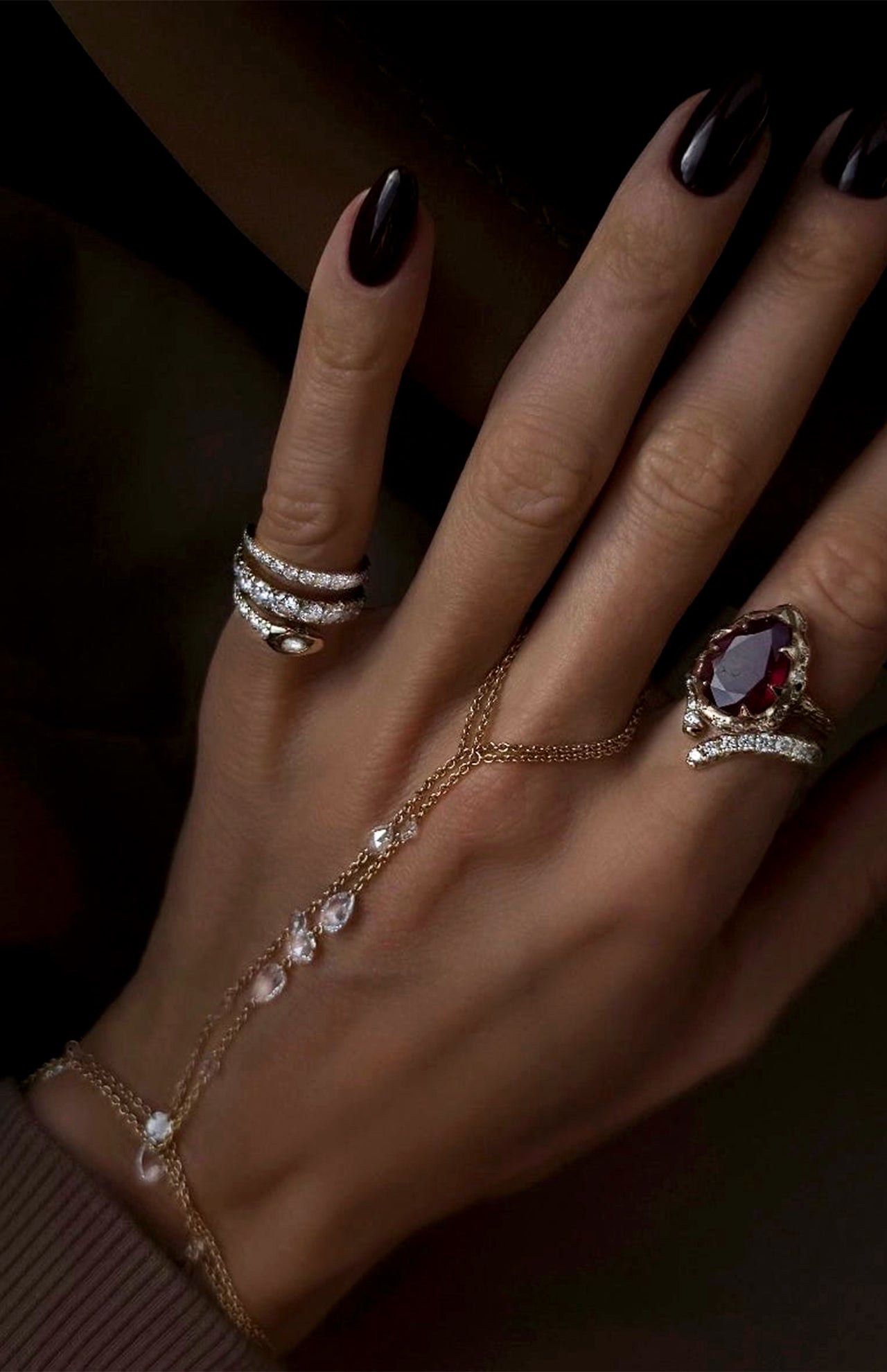 A hand with dark, polished nails wears several rings, including the LOGAN HOLLOWELL French Pave Diamond Tusk Ring and a delicate gold hand chain adorned with small clear stones.