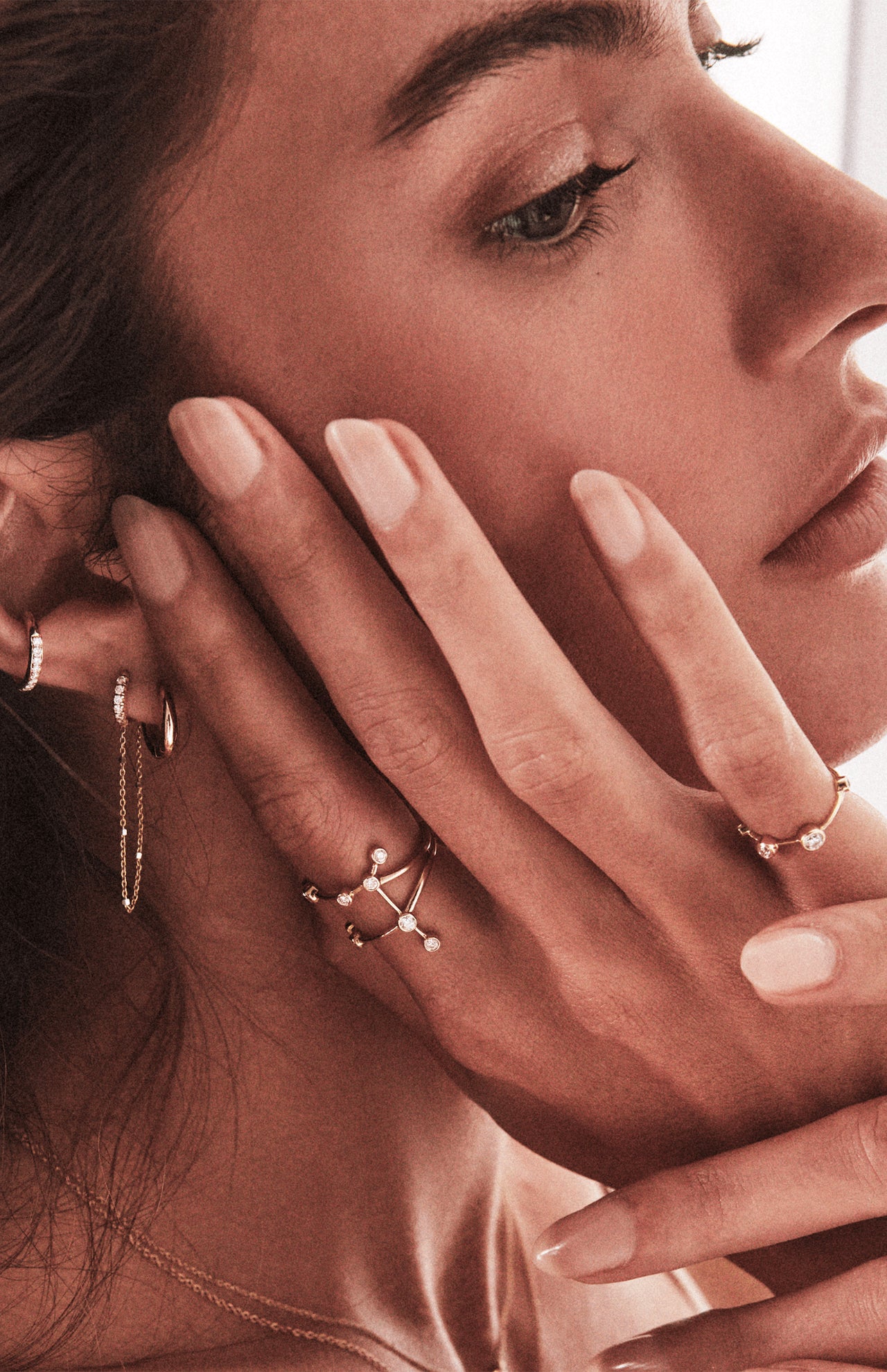 A close-up of a woman’s face and hand highlights LOGAN HOLLOWELL’s Gemini Constellation Ring in yellow gold with diamond accents, paired with gold earrings and her natural manicured nails, showcasing the jewelry's elegance.