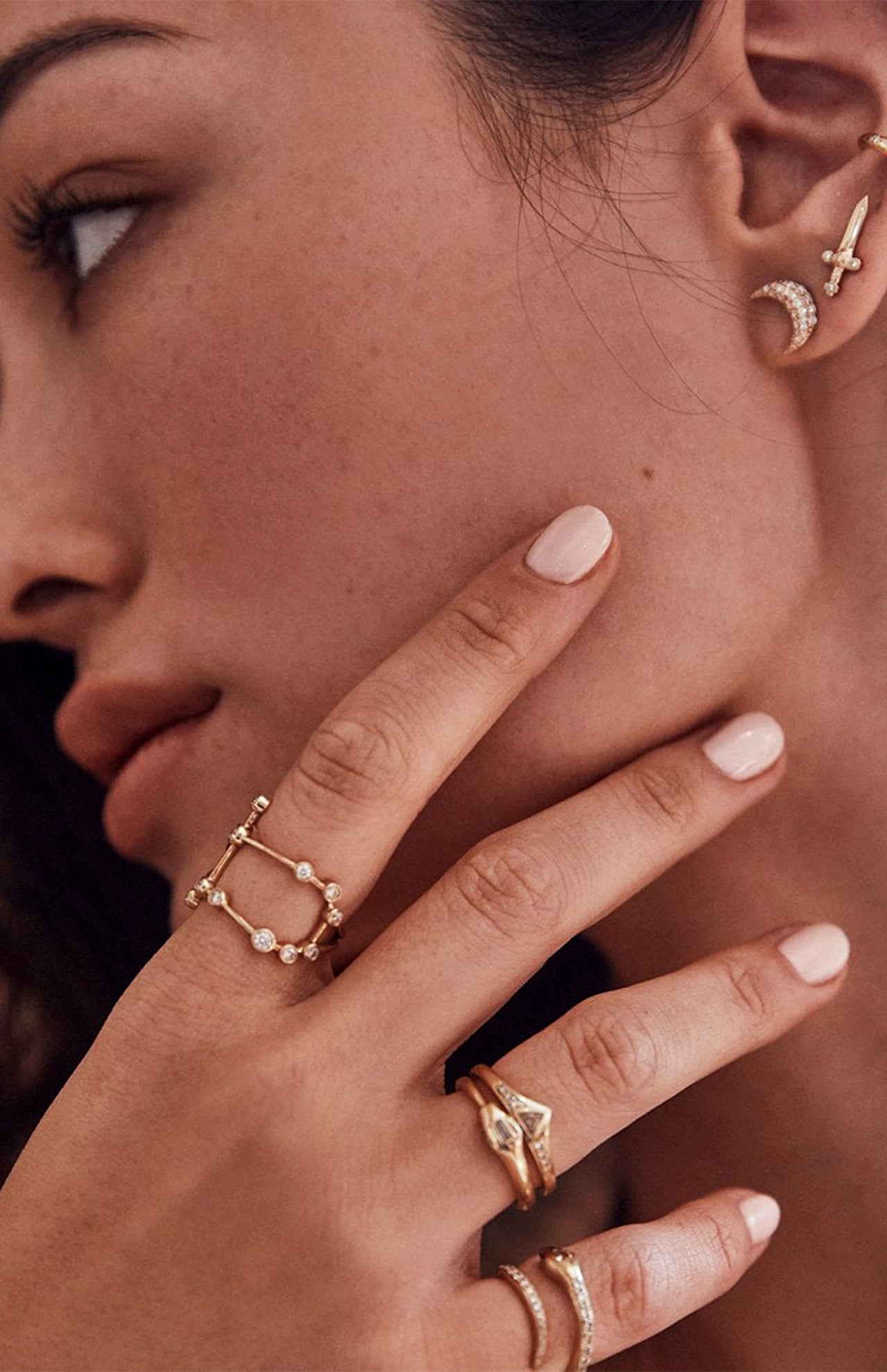 A close-up of a woman’s face and hand with light pink nails, yellow gold rings—including the LOGAN HOLLOWELL Gemini Constellation Ring—and gold crescent moon and star earrings.