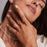 A woman with closed eyes and a serene expression wears layered gold and diamond jewelry, including the LOGAN HOLLOWELL Pisces Diamond Constellation Ring, zodiac necklaces, earrings, a white blouse, and pale pink nail polish.