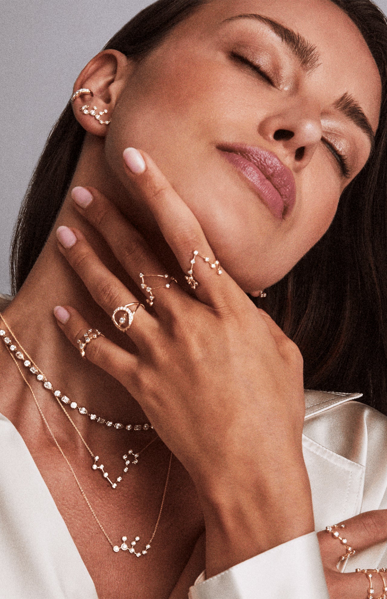 A woman with closed eyes and a serene expression wears layered gold and diamond jewelry, including the LOGAN HOLLOWELL Pisces Diamond Constellation Ring, zodiac necklaces, earrings, a white blouse, and pale pink nail polish.