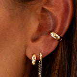 Close-up of a person wearing LOGAN HOLLOWELL's Three Star Set Rounded Diamond Huggie Earrings: stylish 14k gold hoops with diamonds on the lobe and ear, plus a delicate chain. Brown hair is partially visible. Part of our star set collection.