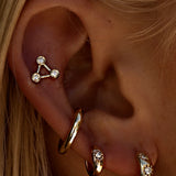 A close-up of a blonde person’s ear with four gold piercings: a triangular gem stud from the Star Set Collection on the upper ear, and three LOGAN HOLLOWELL Three Star Set Rounded Diamond Huggie Earrings on the lower ear.