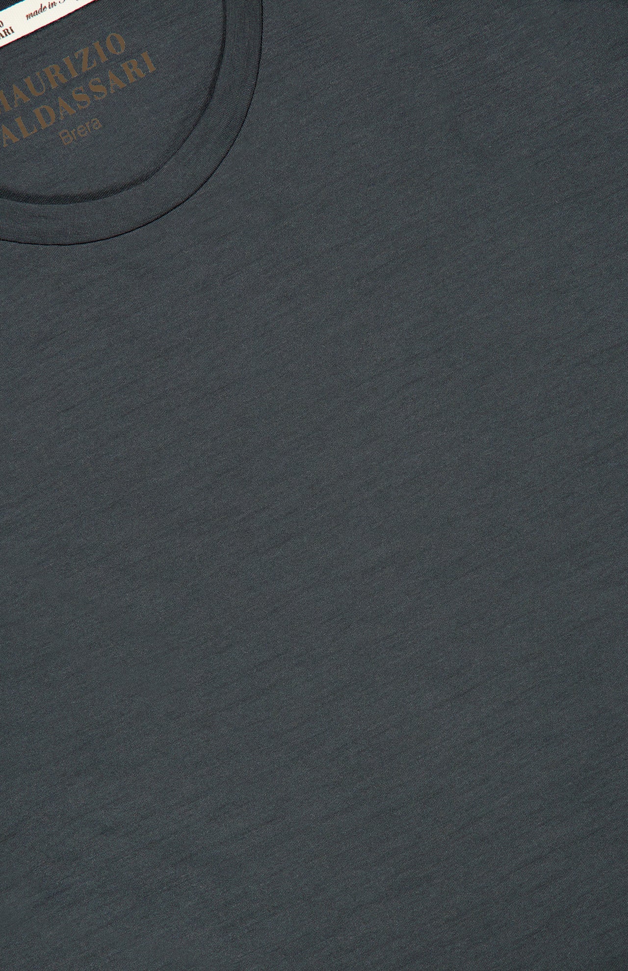 A close-up of a dark gray Linate Short Sleeve T-Shirt by MAURIZIO BALDASSARI, featuring part of the collar and the brand label with gold lettering inside the neckline, highlighting its Made in Italy craftsmanship.