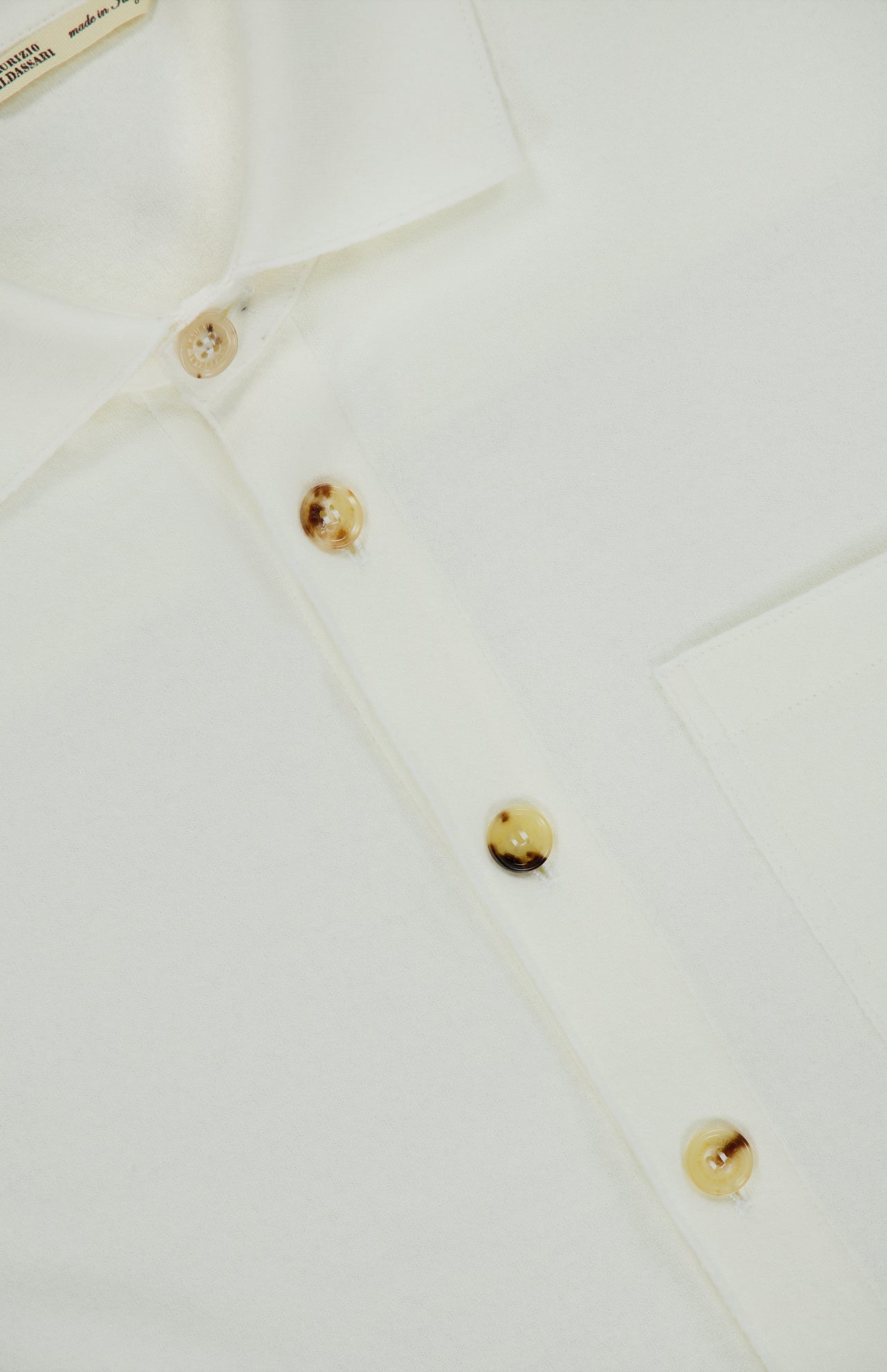 Close-up of the Solferino Wool Overshirt by MAURIZIO BALDASSARI, an off-white Italian-made piece featuring a white collar, chest pocket, and four beige buttons—one fastened at the top and three left unfastened below.