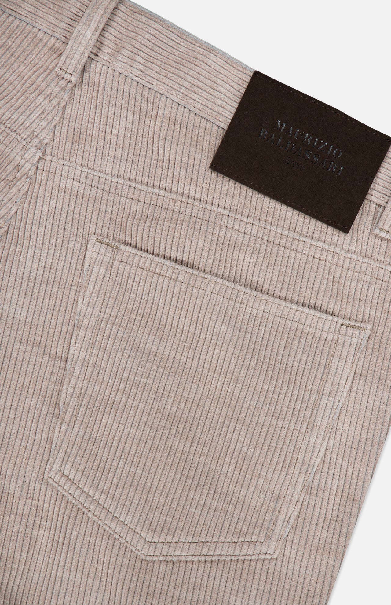 Close-up of the MAURIZIO BALDASSARI Treves Five Pocket Corduroy Pants in taupe, made in Italy, featuring a back pocket and a dark rectangular leather patch near the waistband. The corduroy fabric showcases distinctive vertical ridges.
