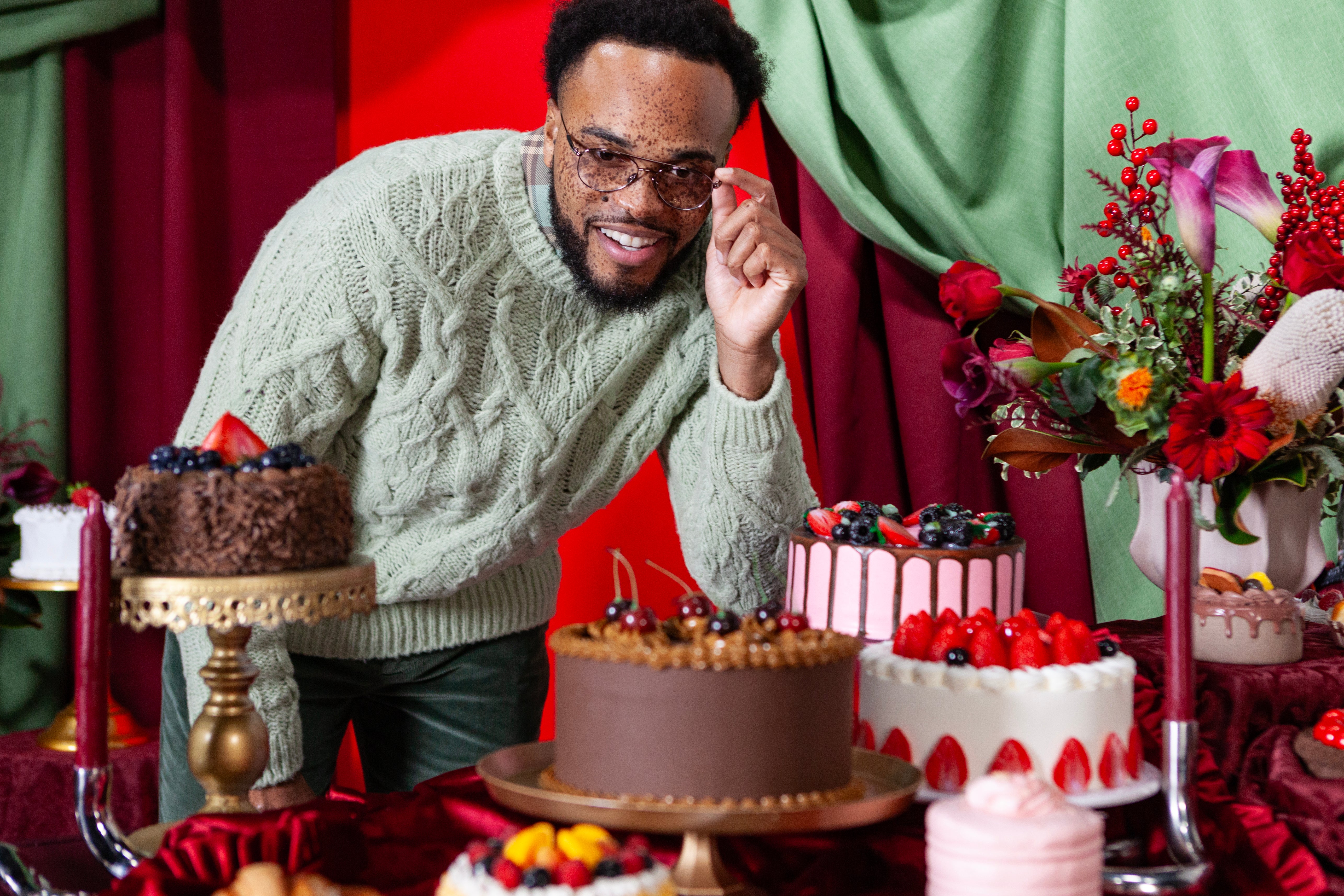 Man in a green sweater standing behind a table with cakes and flowers in a festive setting.