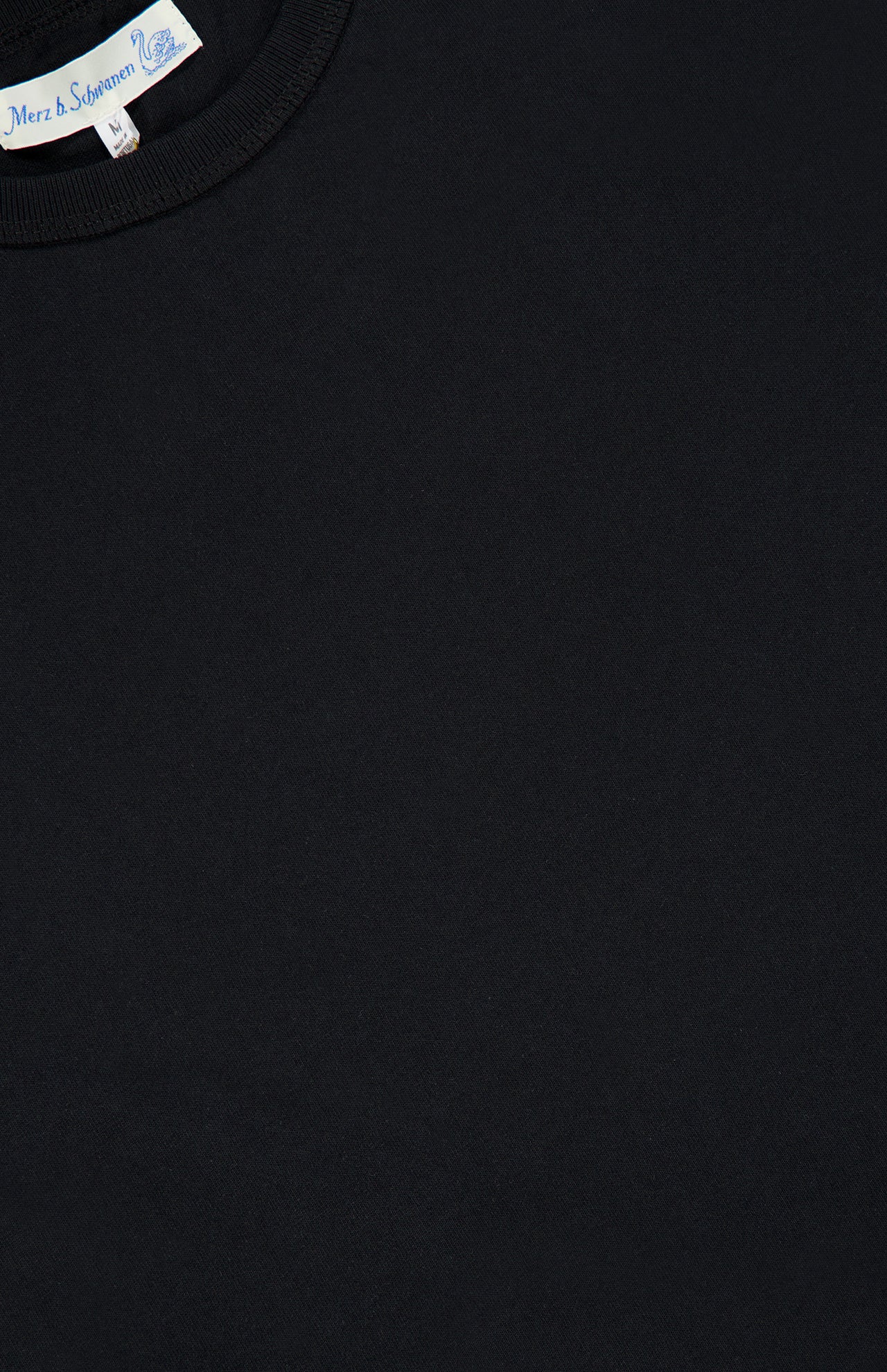 Close-up of the Classic Jersey T-shirt by Merz B. Schwanen in plain black mid-weight jersey, showcasing a visible neck collar and a partially visible white label with the brand’s name, emphasizing its heritage craftsmanship.