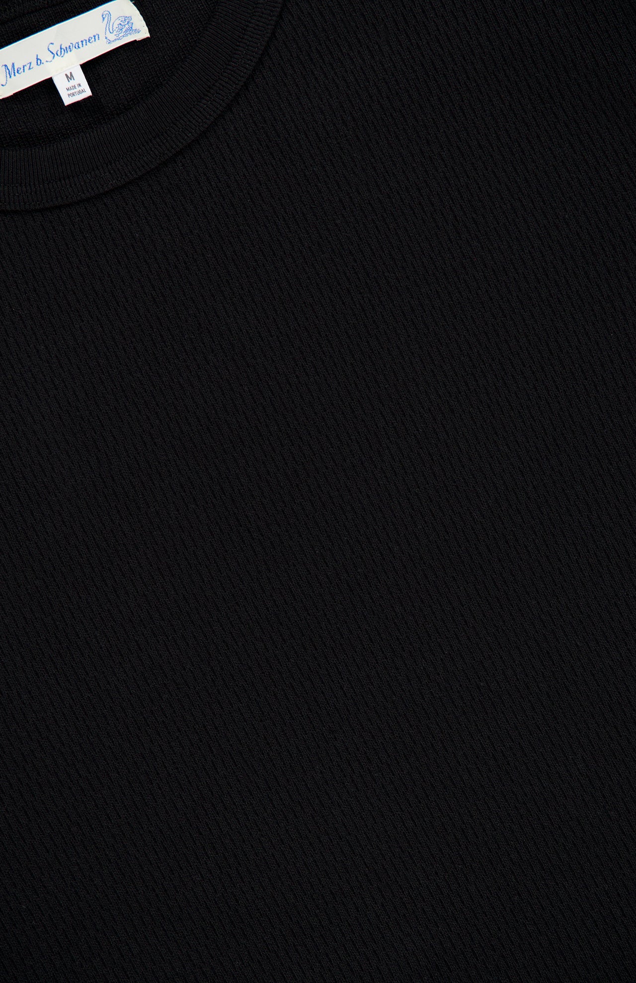 Close-up of the Merz B. Schwanen Cotton Thermal Long Sleeve T-shirt in black, showcasing a textured crewneck and a visible white collar tag displaying the brand name and size M for medium. Made from organic cotton, this timeless piece offers exceptional comfort and quality.
