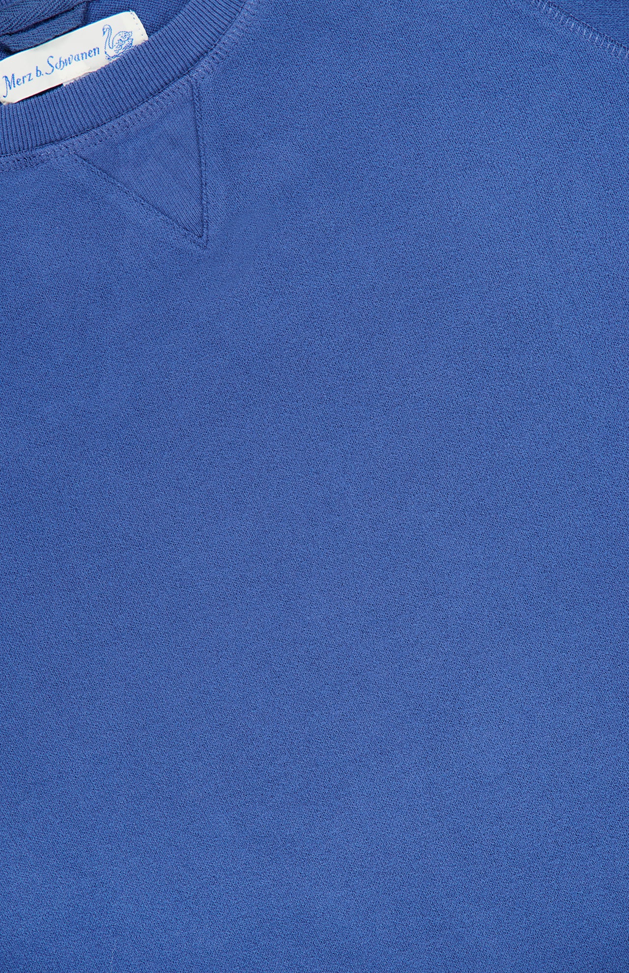 A close-up of the Merz B. Schwanen Light Weight Crewneck Sweatshirt in blue, made from organic cotton, features a ribbed collar, V-stitch detail, and part of a white tag with text.