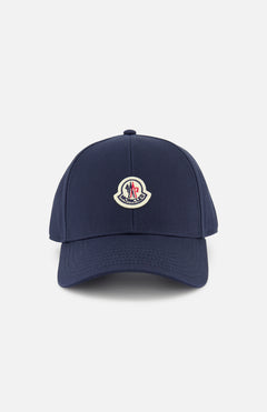 The MONCLER Logo Baseball Cap in navy blue features a curved brim, iconic front logo patch, and adjustable back strap, shown against a plain white background.