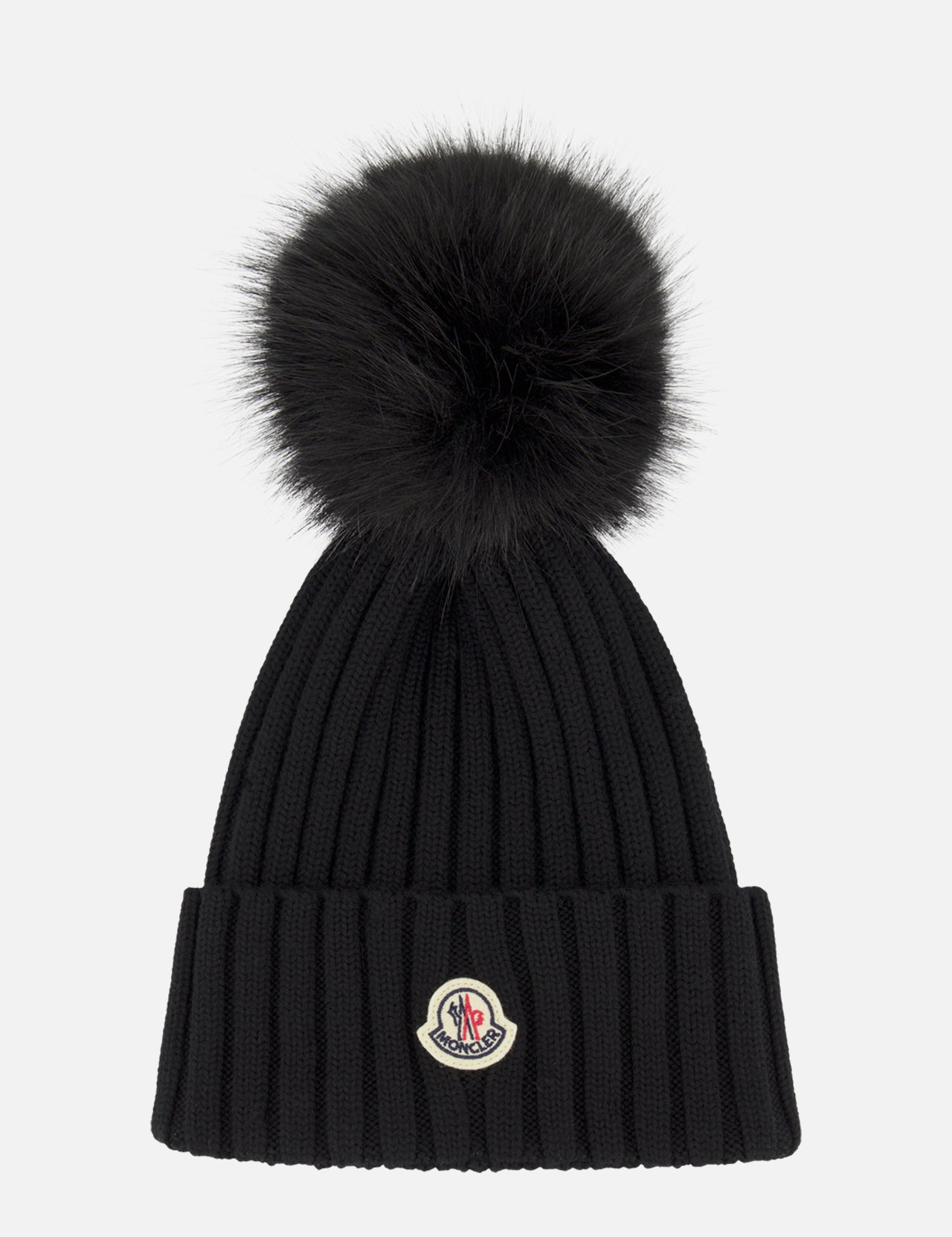 The MONCLER Faux Fur Pom Pom Beanie is a black wool rib-knit hat with a large black faux fur pom-pom and an iconic logo patch on the folded cuff, shown against a plain white background.