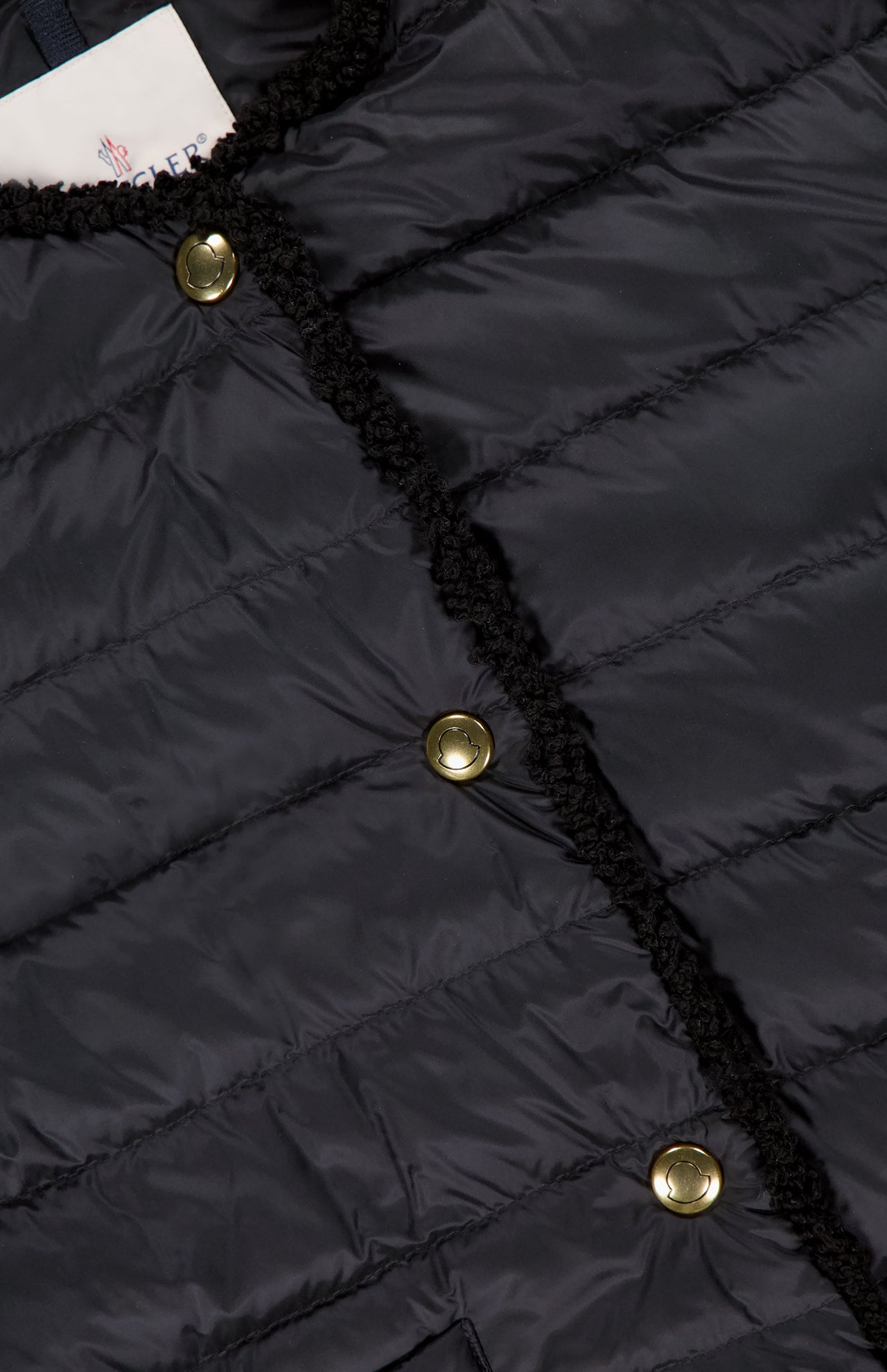 Close-up of the MONCLER Fleurs Bouclé Trim Jacket, featuring a dark quilted texture, water-repellent fabric, three shiny gold buttons, black trim along the front opening, and a partially visible clothing label.